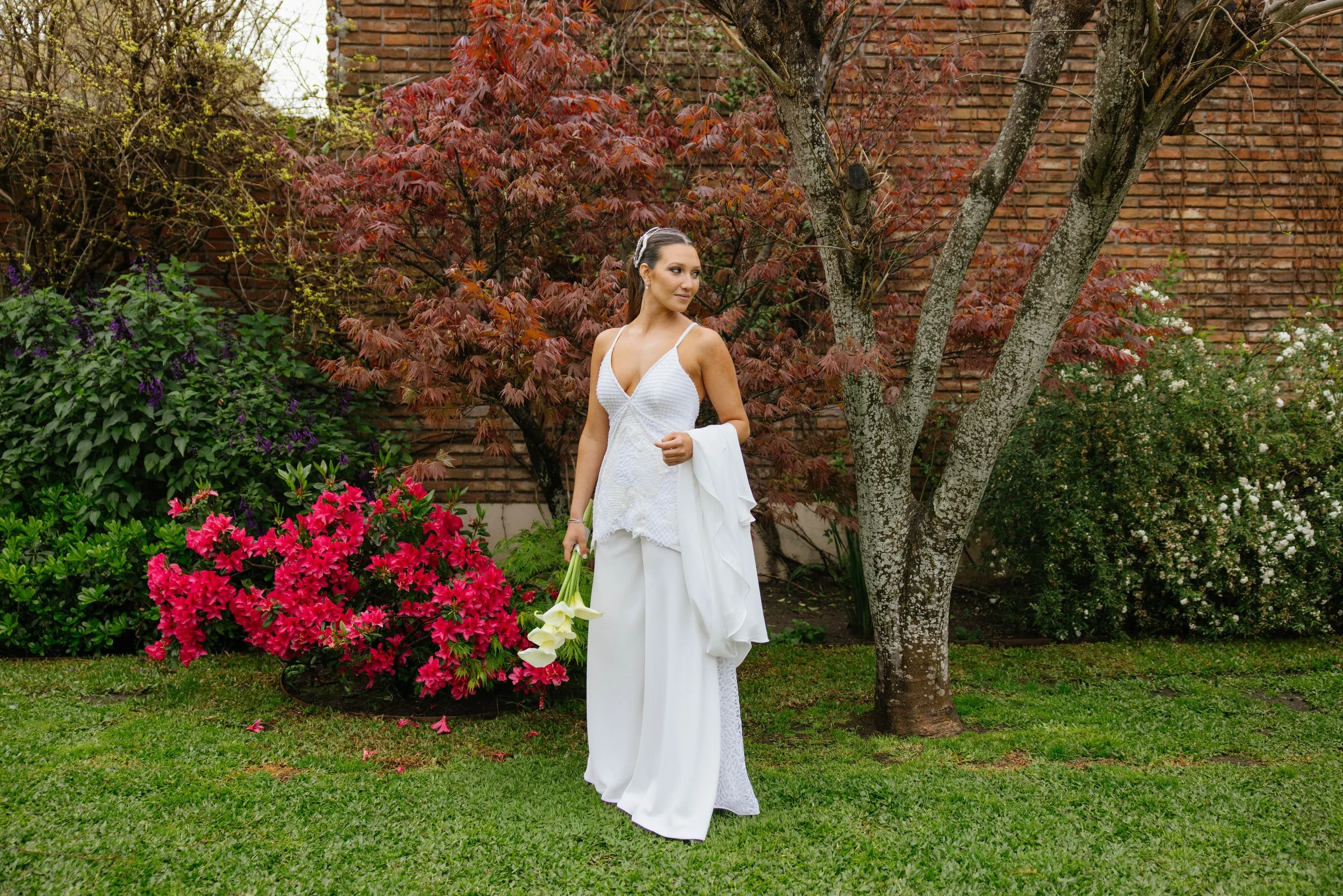 Mujer vestida con un vestido blanco, sosteniendo un ramo de flores blancas, de pie en un jardín con arbustos de flores rosadas y blancas, y un árbol con ramas delgadas y un muro de ladrillos en el fondo.