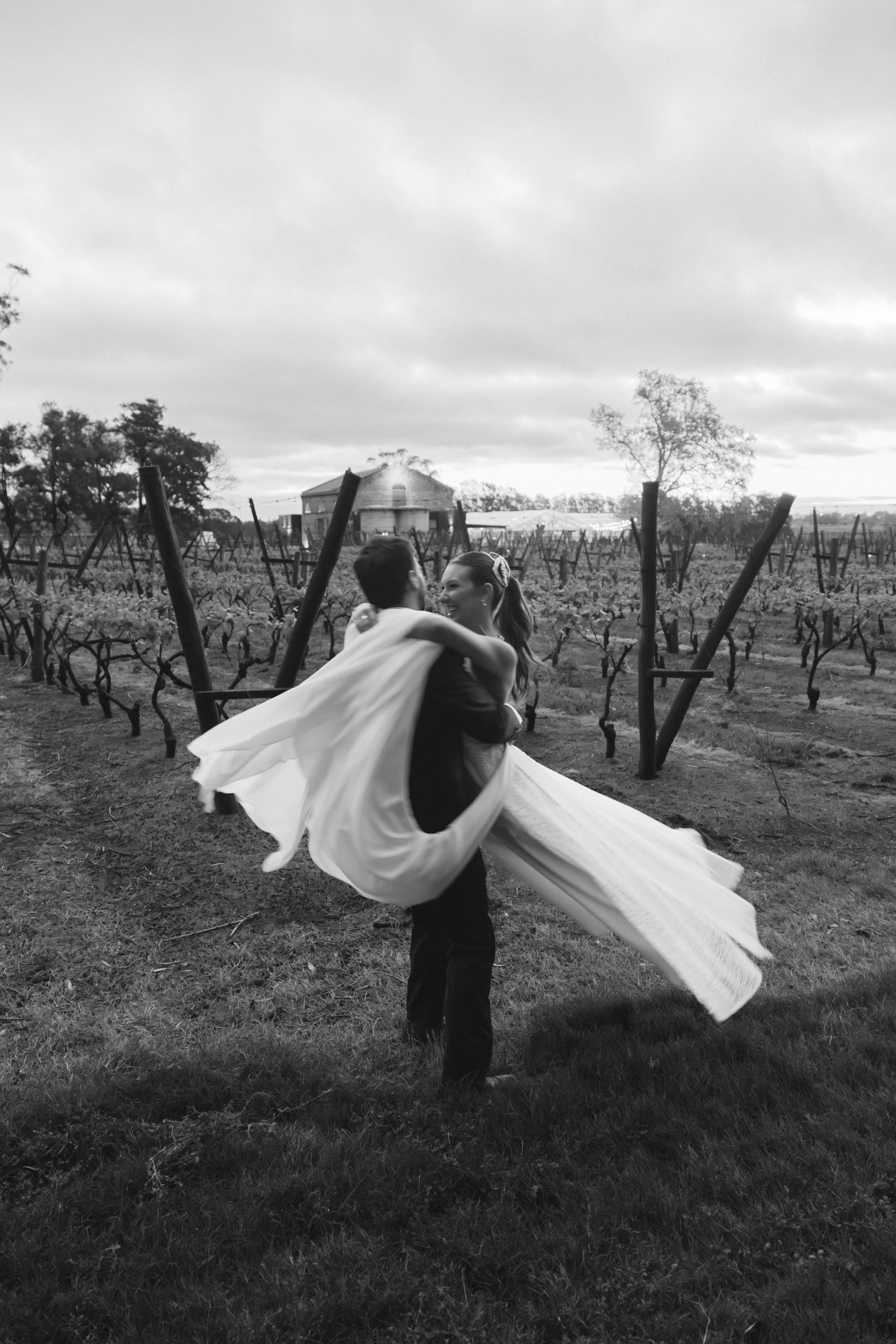 Pareja bailando en un viñedo, la mujer lleva un vestido largo y el hombre un traje oscuro, en un día nublado.