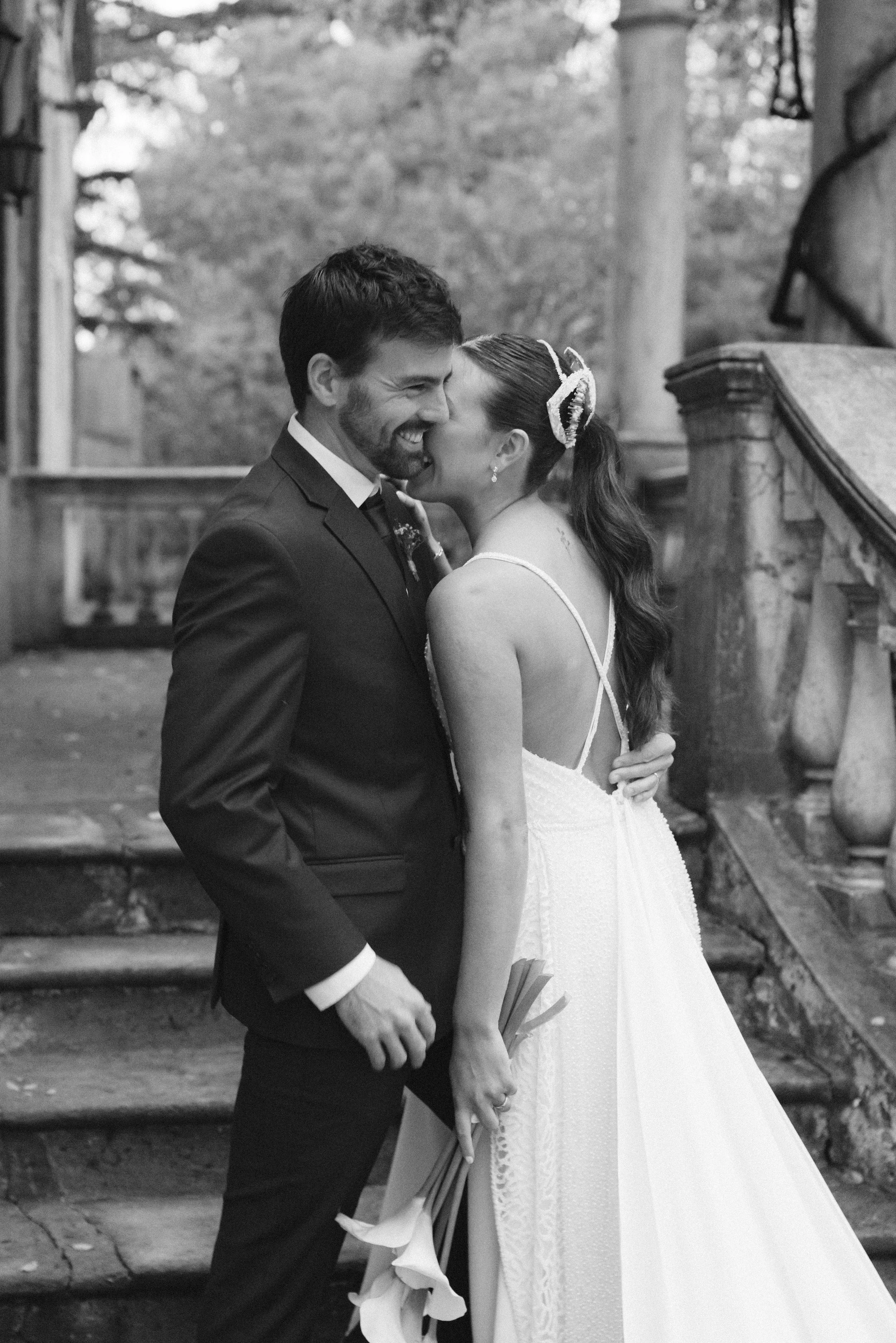 Una pareja se besa frente a unas escaleras, ella lleva un vestido de novia y él un traje oscuro. Ambos parecen felices, rodeados de un entorno natural con árboles.