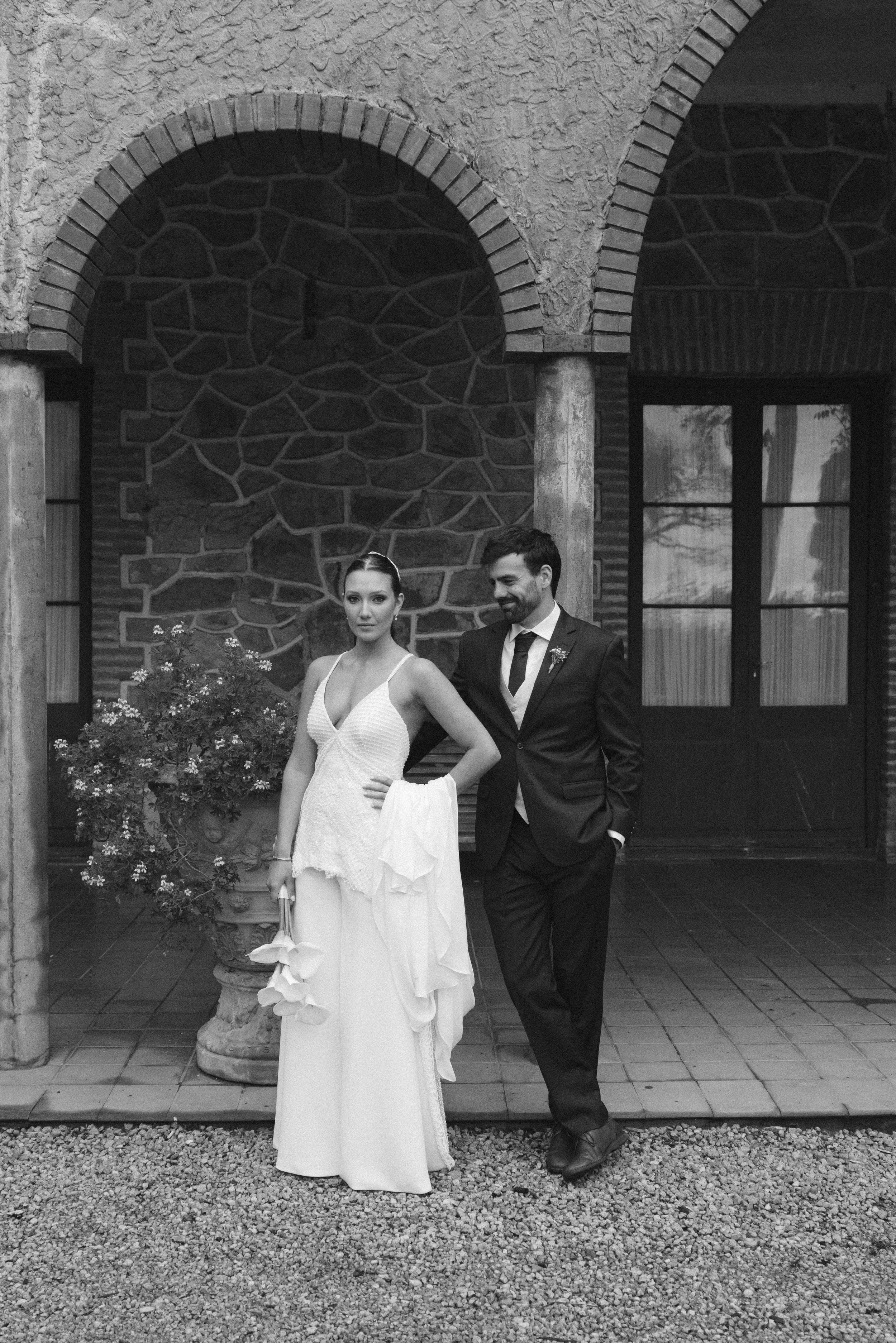 Una pareja de novios en boda, la mujer lleva vestido de novia y el hombre saco, en un fondo de muro de piedra y ventanas.