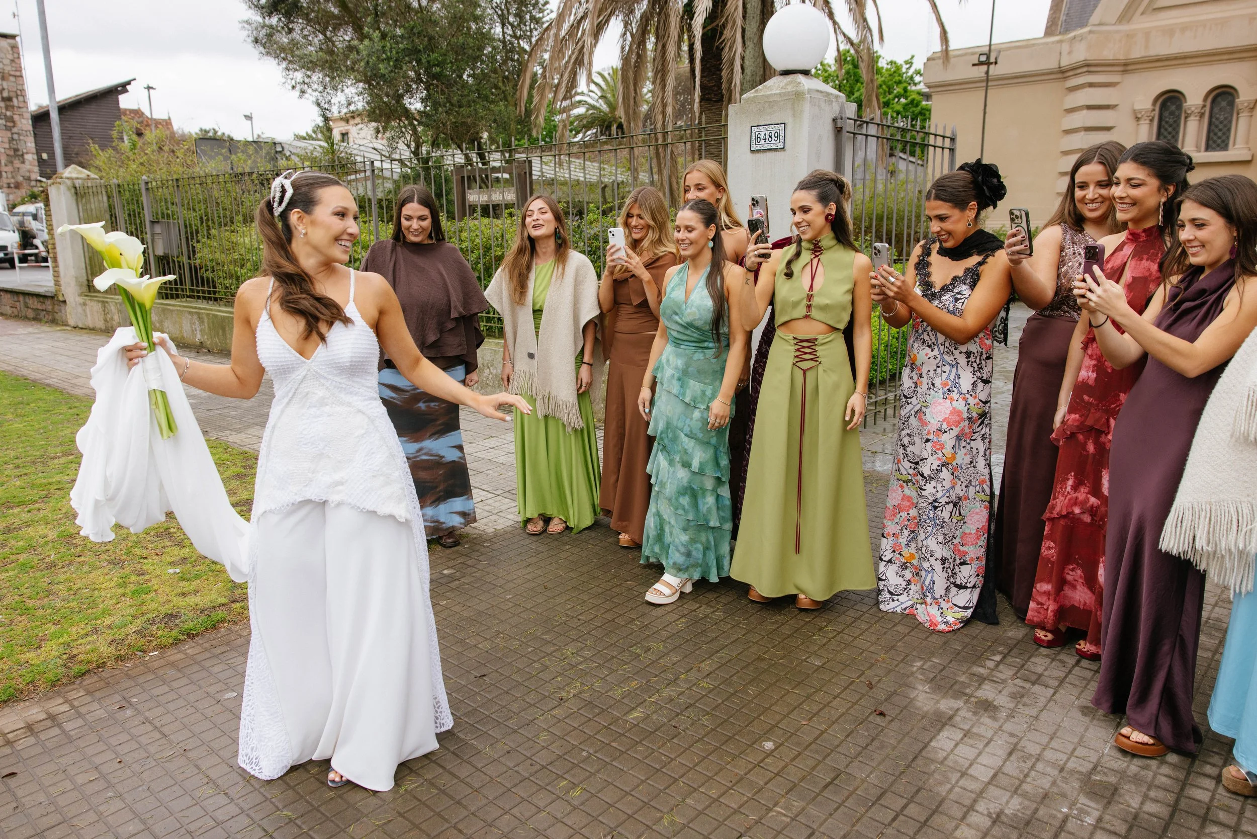 Mujeres con vestidos largos y coloridos, una de ellas con vestido de novia, rodeadas de otras mujeres, en una ceremonia al aire libre en un espacio urbano con árboles y edificios, algunas tomando fotos con teléfonos móviles.