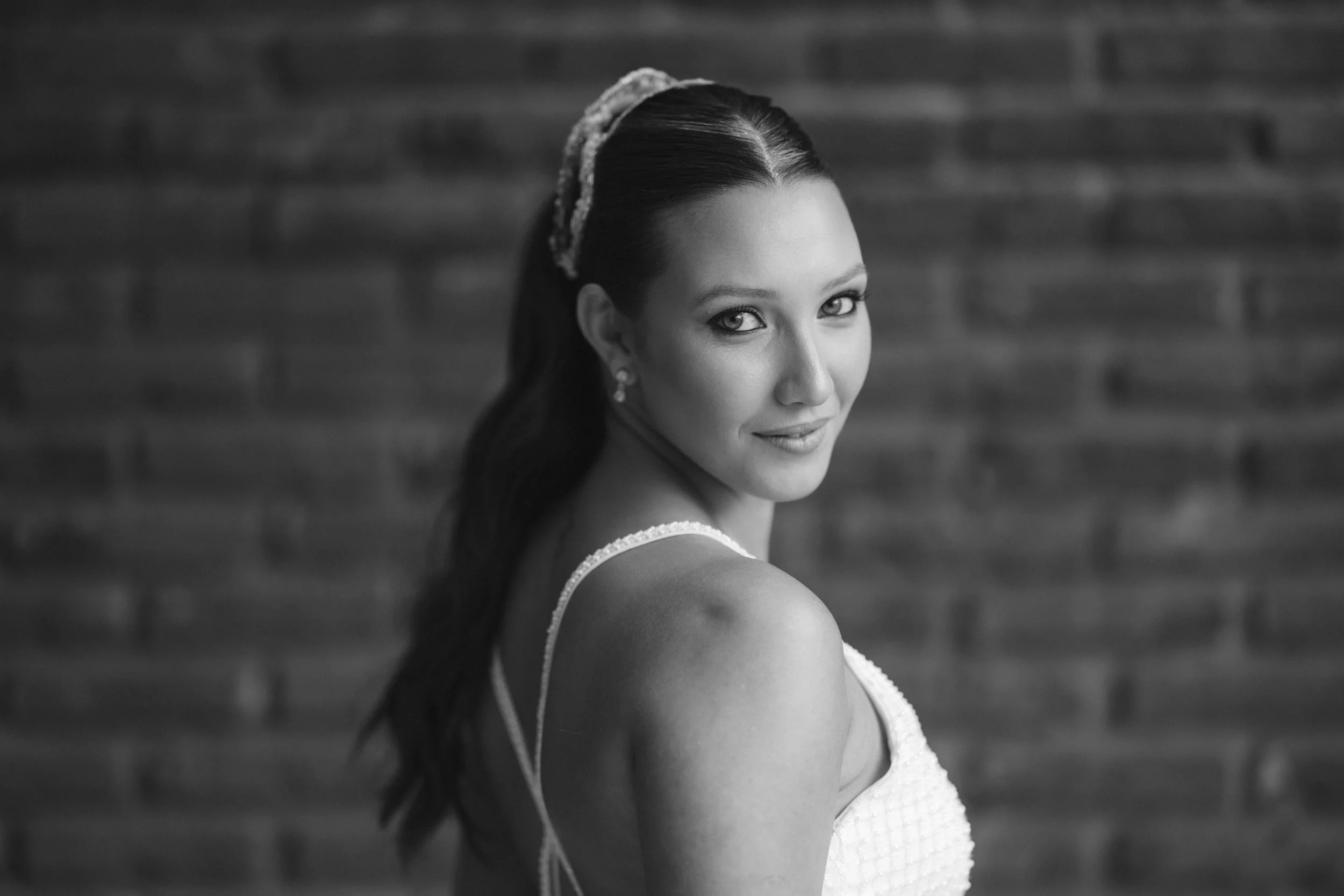 Retrato en blanco y negro de una mujer con cabello oscuro, peinada con una raya en medio, y con una diadema decorativa. Ella está sonriendo suavemente y lleva un vestido blanco con tirantes. La mujer mira hacia la cámara, con fondo de pared de ladril