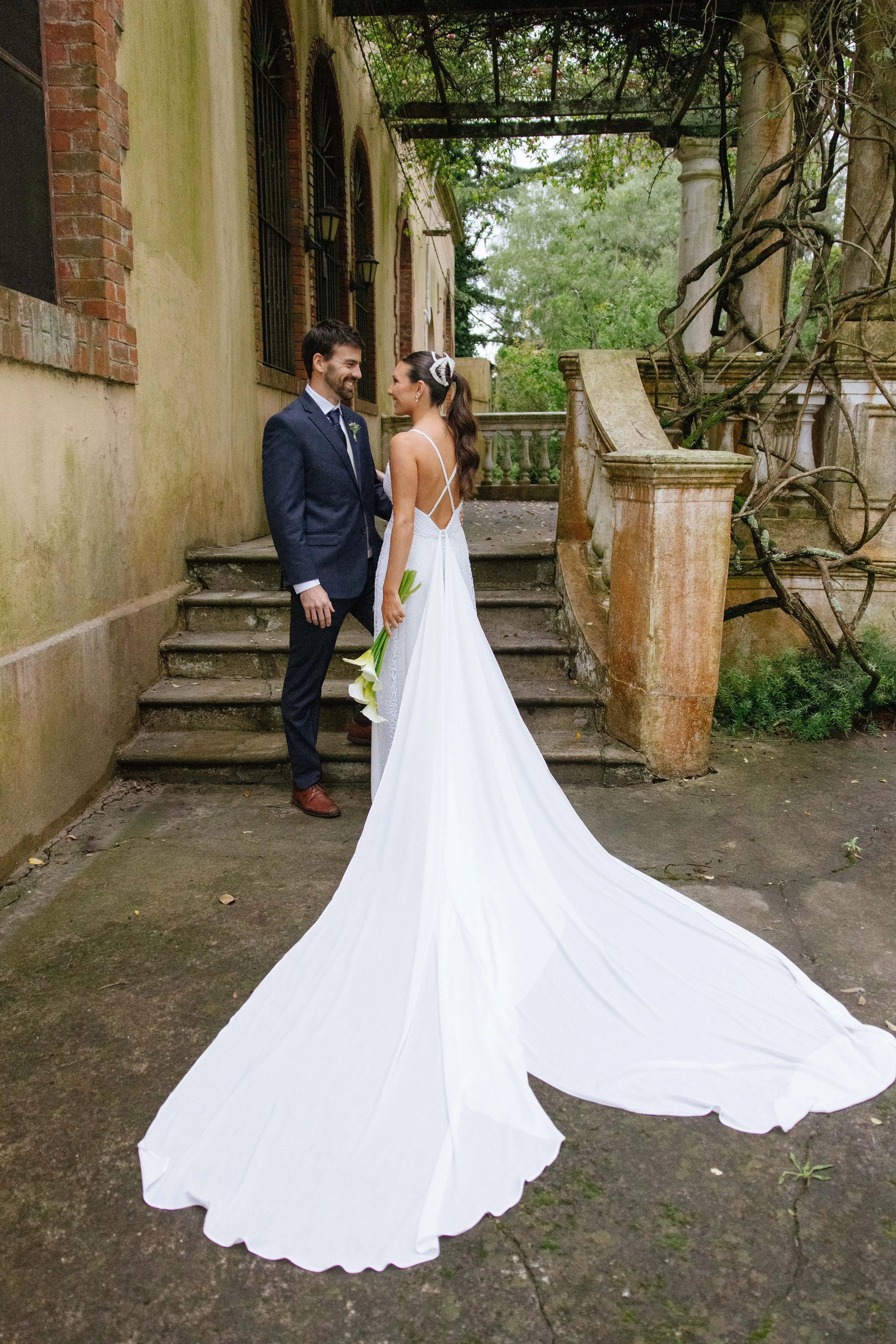 Pareja de recién casados, hombre con traje oscuro y mujer con vestido de novia y bouquet, de pie en un pasaje con escalones y columnas, entorno con vegetación y estructura envejecida.