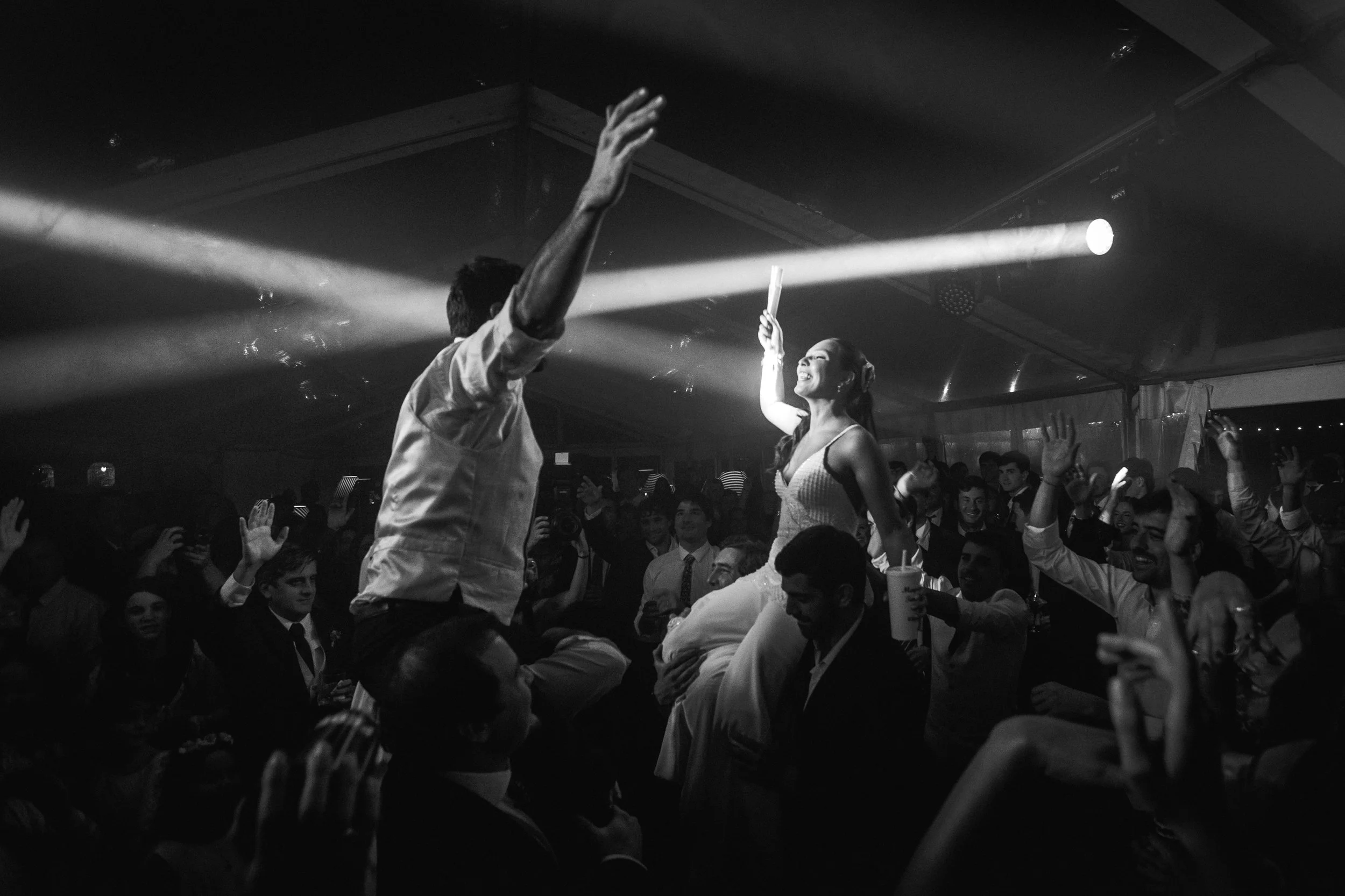 Pareja bailando en una fiesta, rodeada de muchas personas que también disfrutan y levantan las manos, en un ambiente animado y festivo.