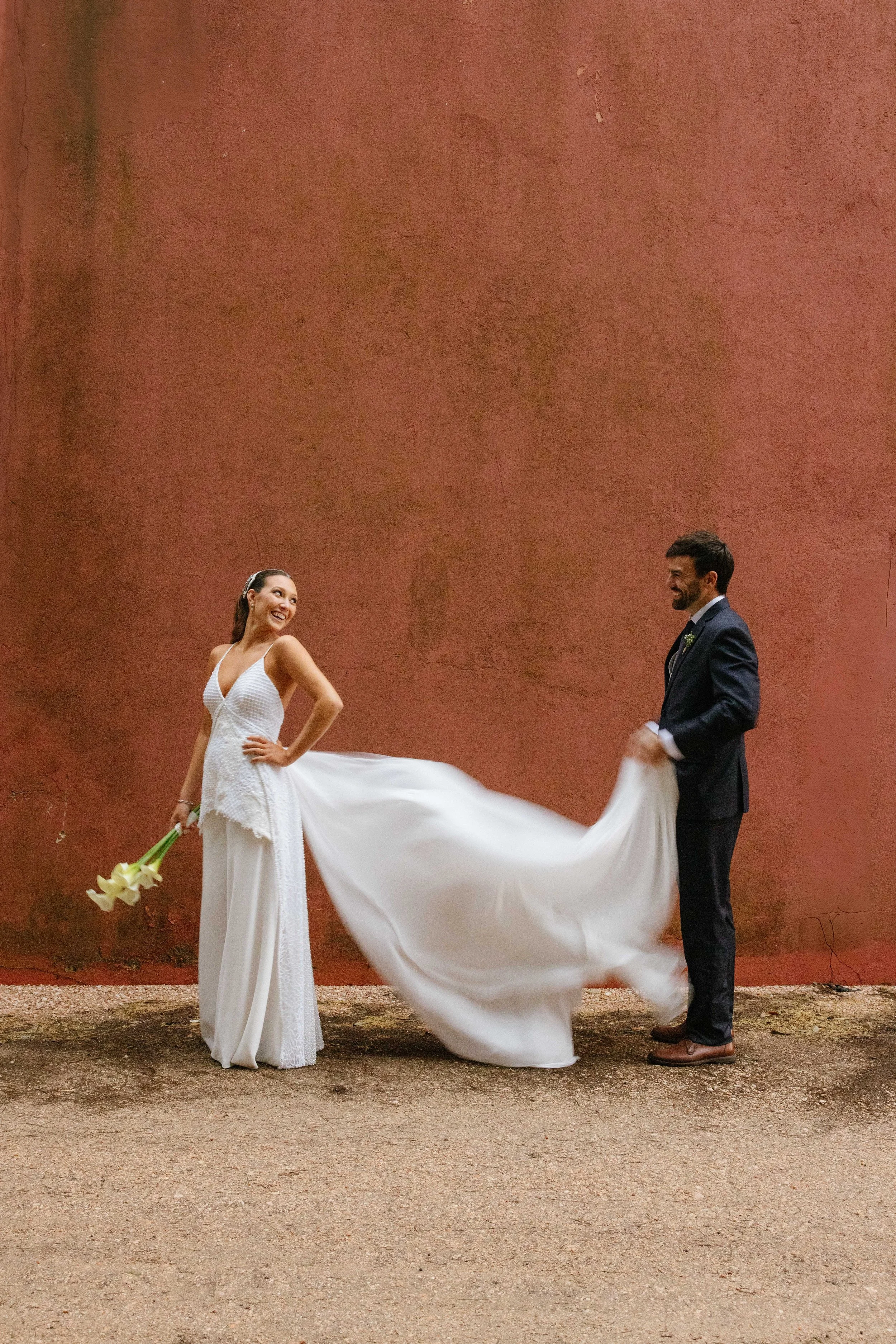 Pareja de novios bailando en una boda frente a una pared de color terracota, la mujer lleva un vestido de novia y sostiene un ramo de lirios, el hombre viste un traje oscuro y ambos sonríen.