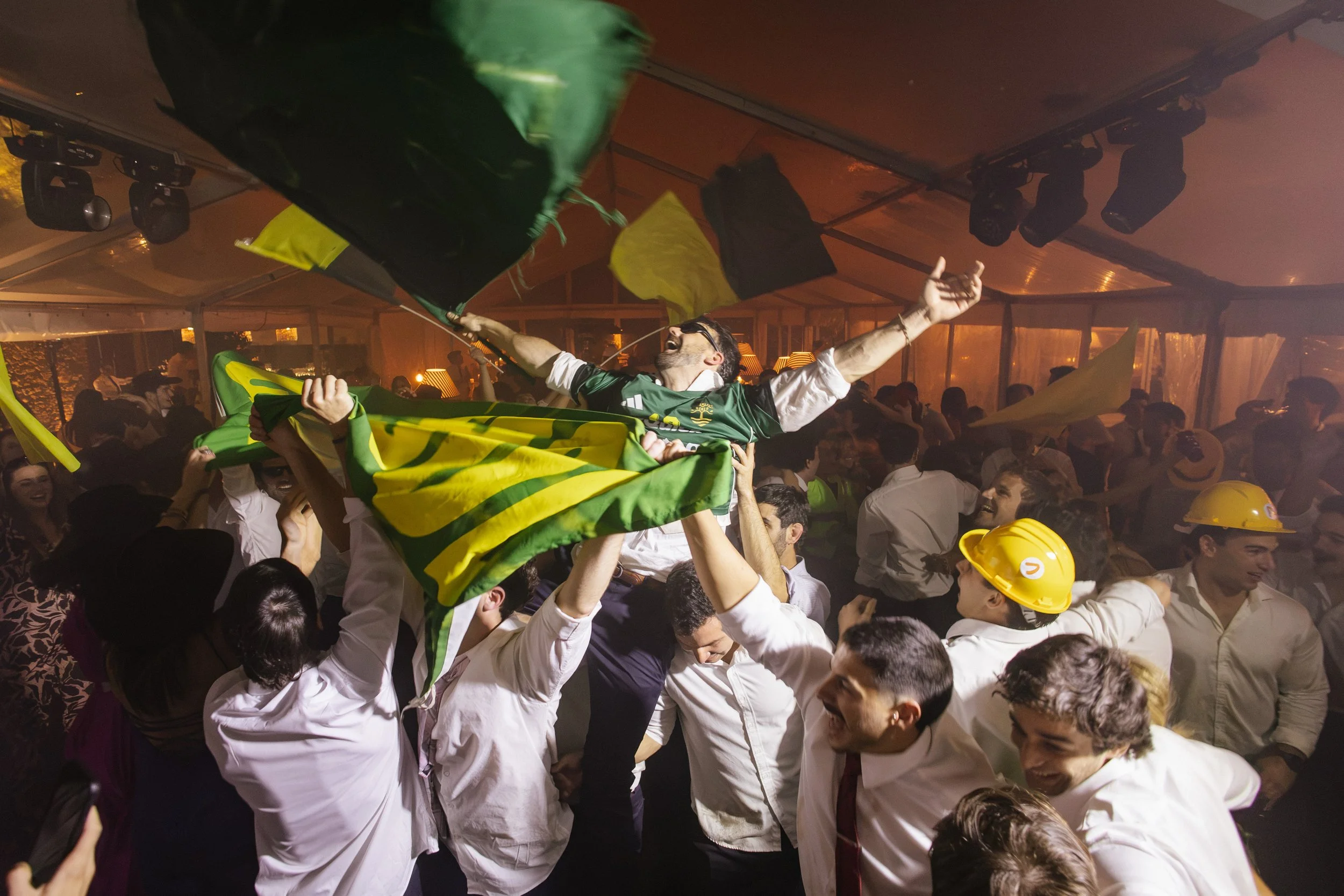 Un grupo de personas en una celebración, levantando a una persona en el aire que lleva una bandera verde y amarilla y ropa deportiva, en un entorno festivo con ambiente de fiesta y humo en el fondo.