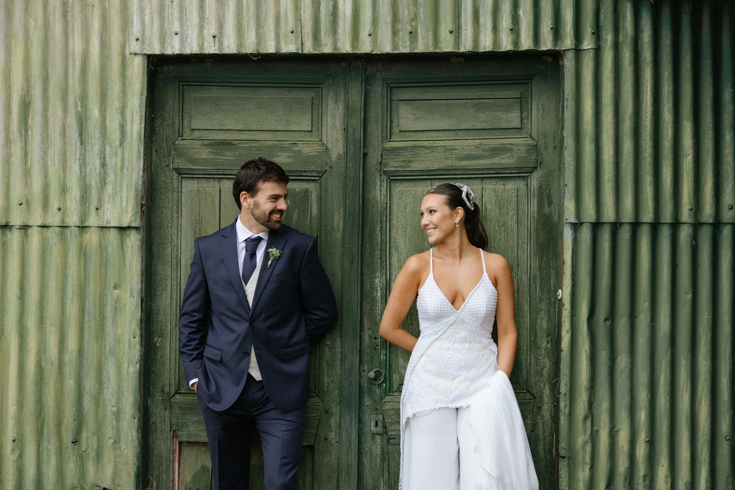 Pareja de boda, hombre con traje azul oscuro y mujer con vestido blanco, sonriendo frente a una puerta de madera verde desgastada.
