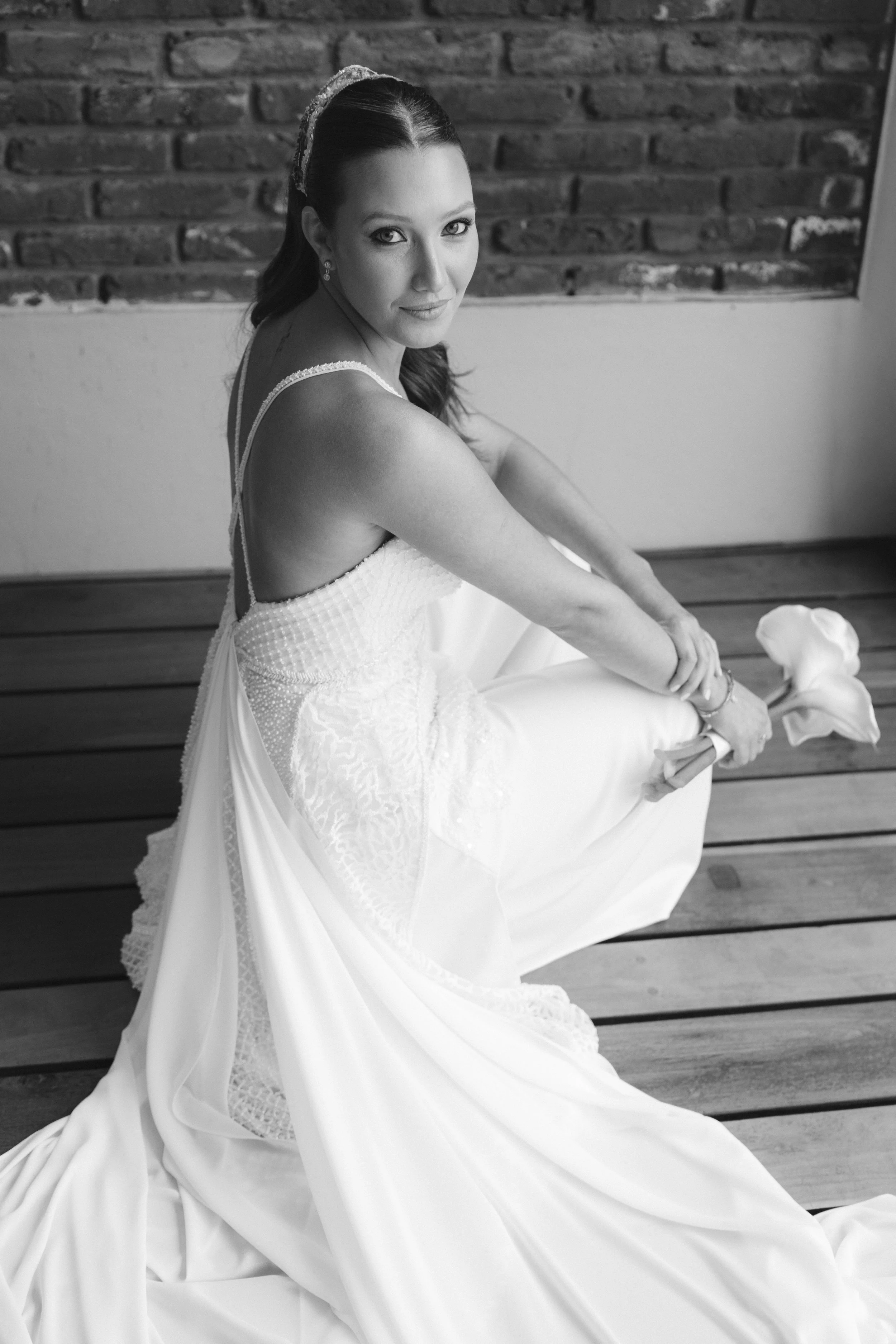 Mujer con vestido de novia sentada sobre un banco de madera, mirando hacia la cámara, en un ambiente interior con pared de ladrillos y piso de madera.