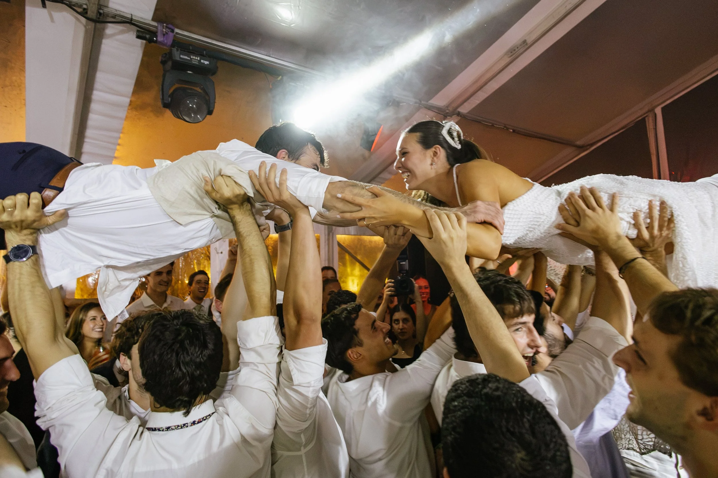 Personas participando en una fiesta, sosteniendo a una pareja que está siendo levantada por las manos del grupo, en un ambiente festivo con muchas sonrisas.