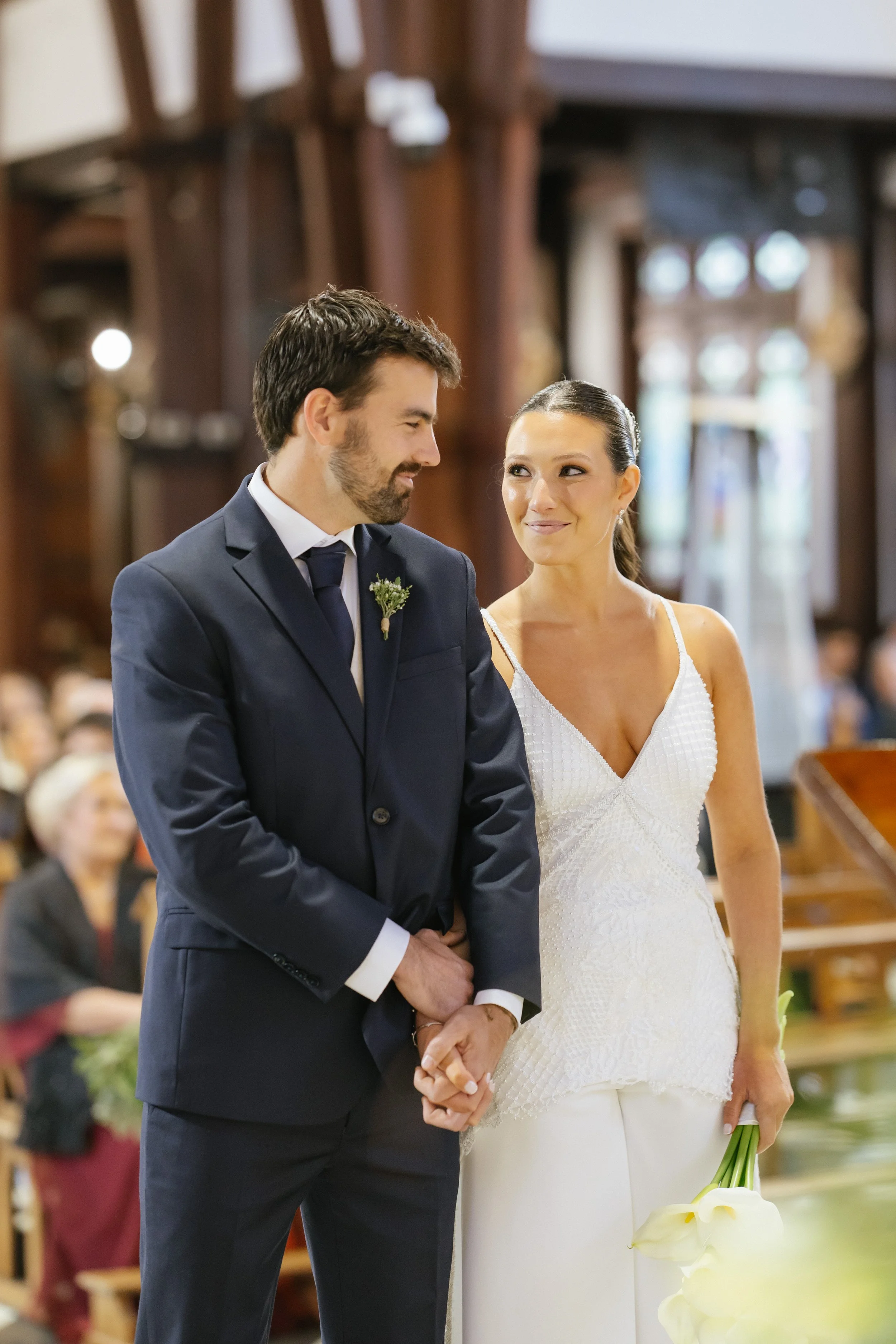 Una pareja en una ceremonia religiosa de matrimonio, sosteniéndose de las manos, con invitados al fondo en una iglesia de madera decorada.