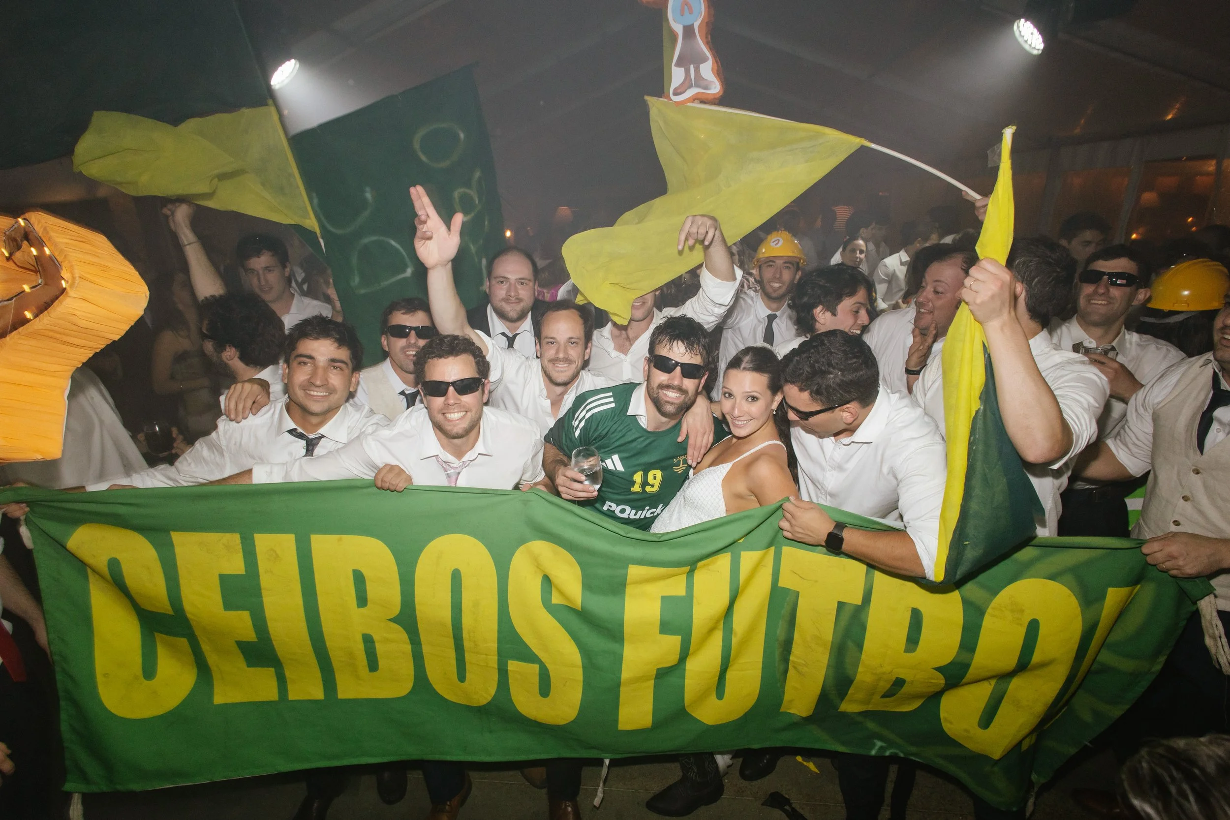 Grupo de personas celebrando con banderas y camisetas de colores en un evento, algunos con gafas de sol y otros usando cascos amarillos.