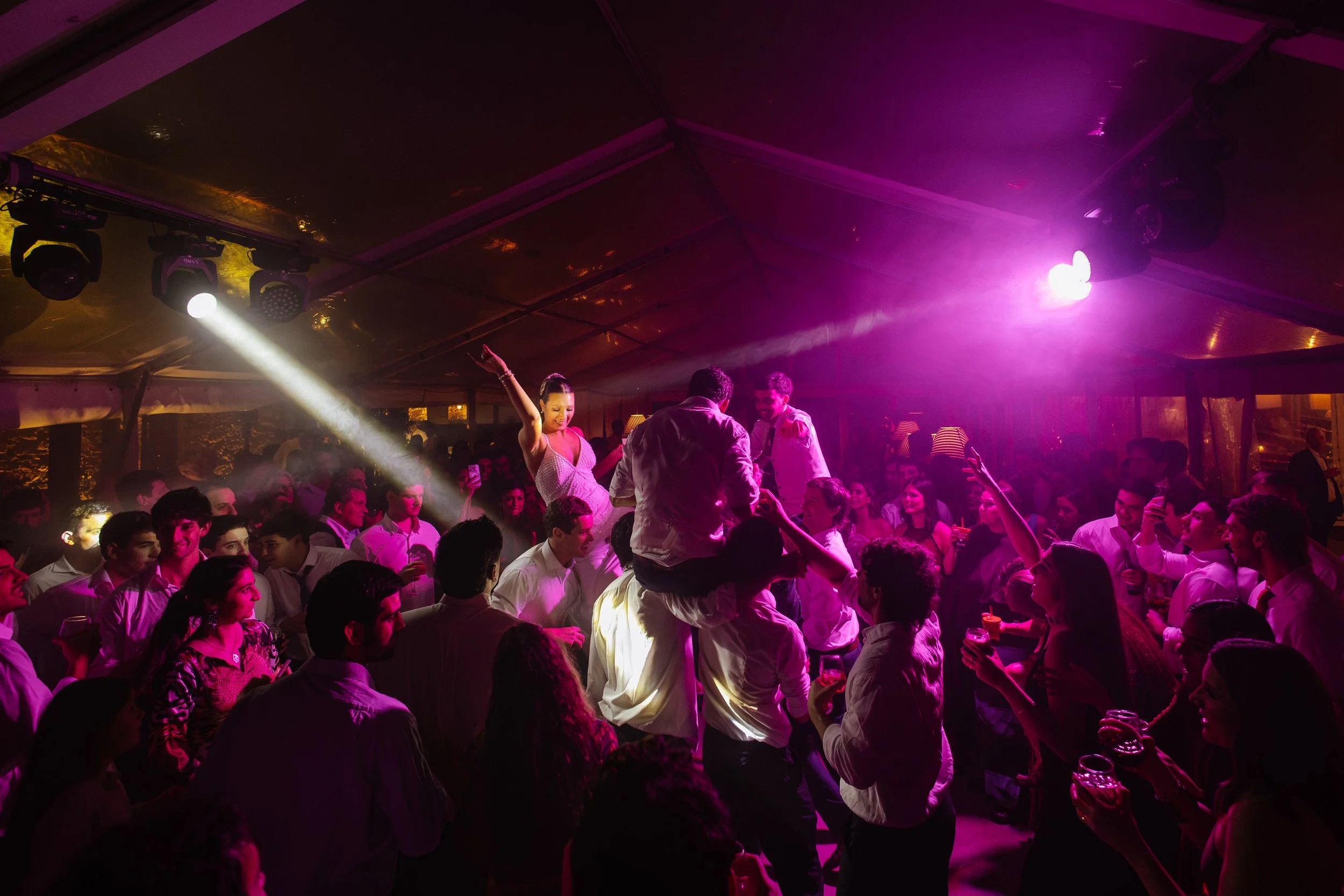 Un grupo de personas bailando en una fiesta con iluminación rosa y morada, con algunos asistentes en el centro de la pista y otros en las mesas