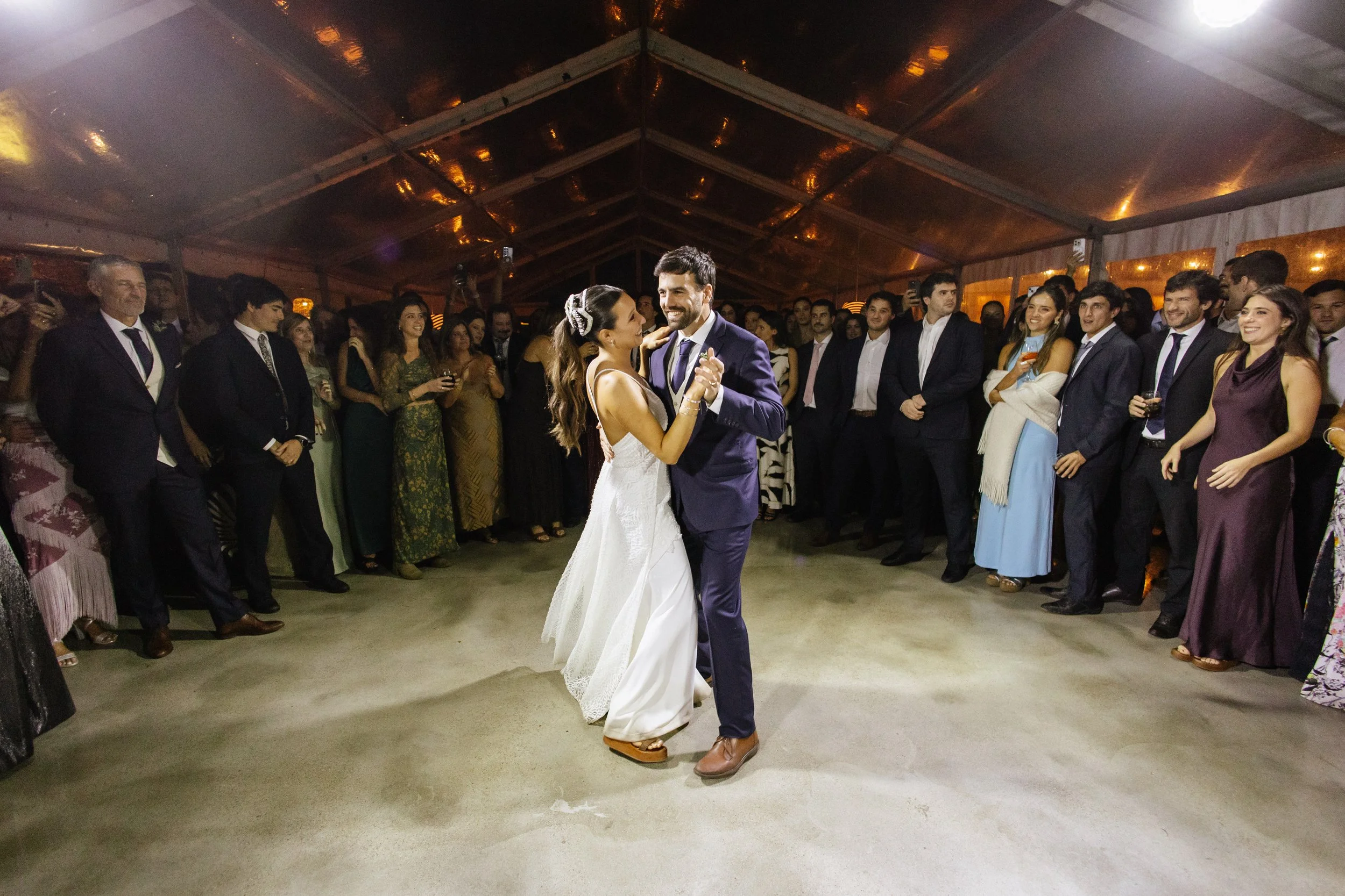 Pareja bailando en un salón de boda con invitados observando, ambiente cálido y elegante bajo un techo de madera y luces. La mujer lleva vestido de novia y el hombre traje formal.