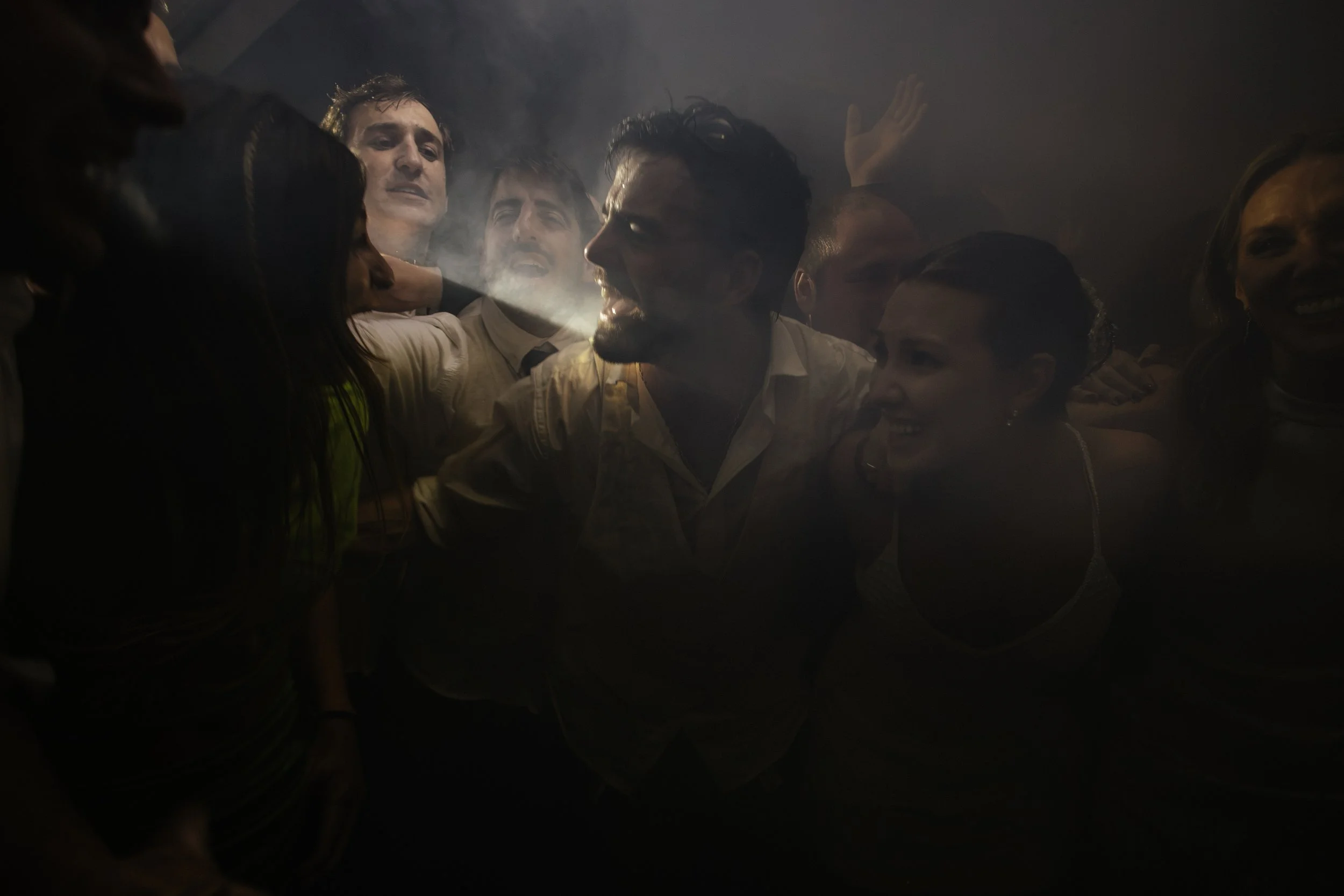 Grupo de personas en una fiesta, algunas riendo y otras con expresiones de emoción, en un ambiente oscuro con luz suave y humo.