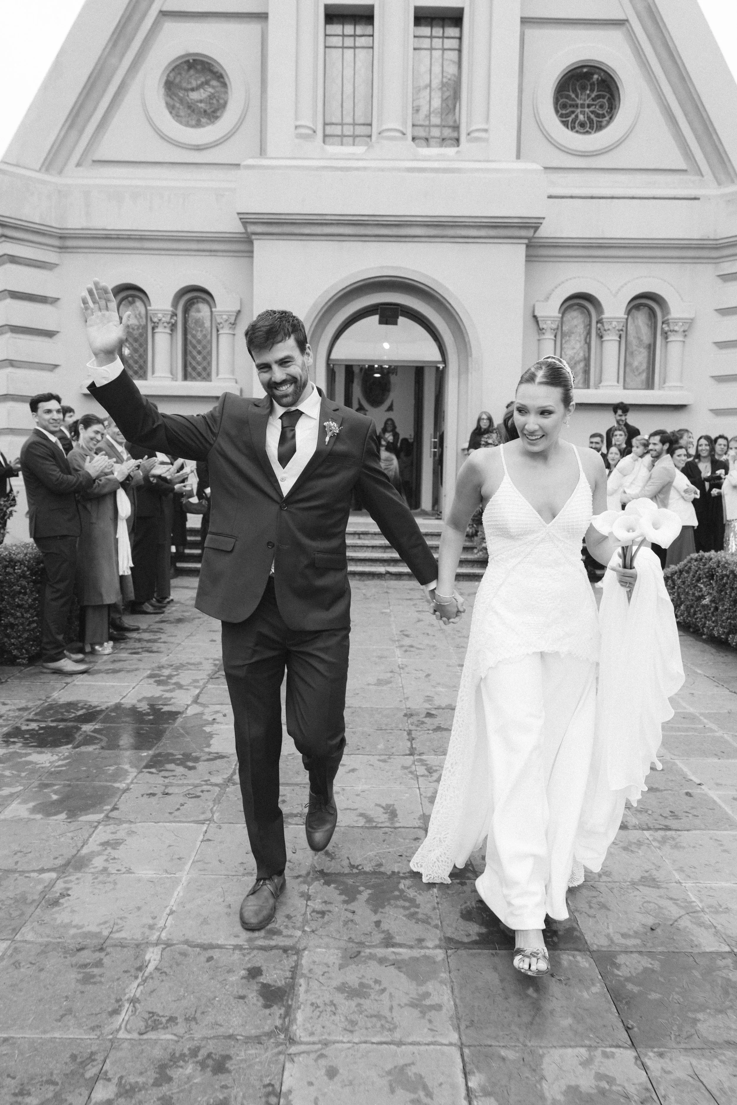 Pareja de recién casados, ella lleva vestido blanco y él traje oscuro, caminando felices frente a un edificio y rodeados de invitados.