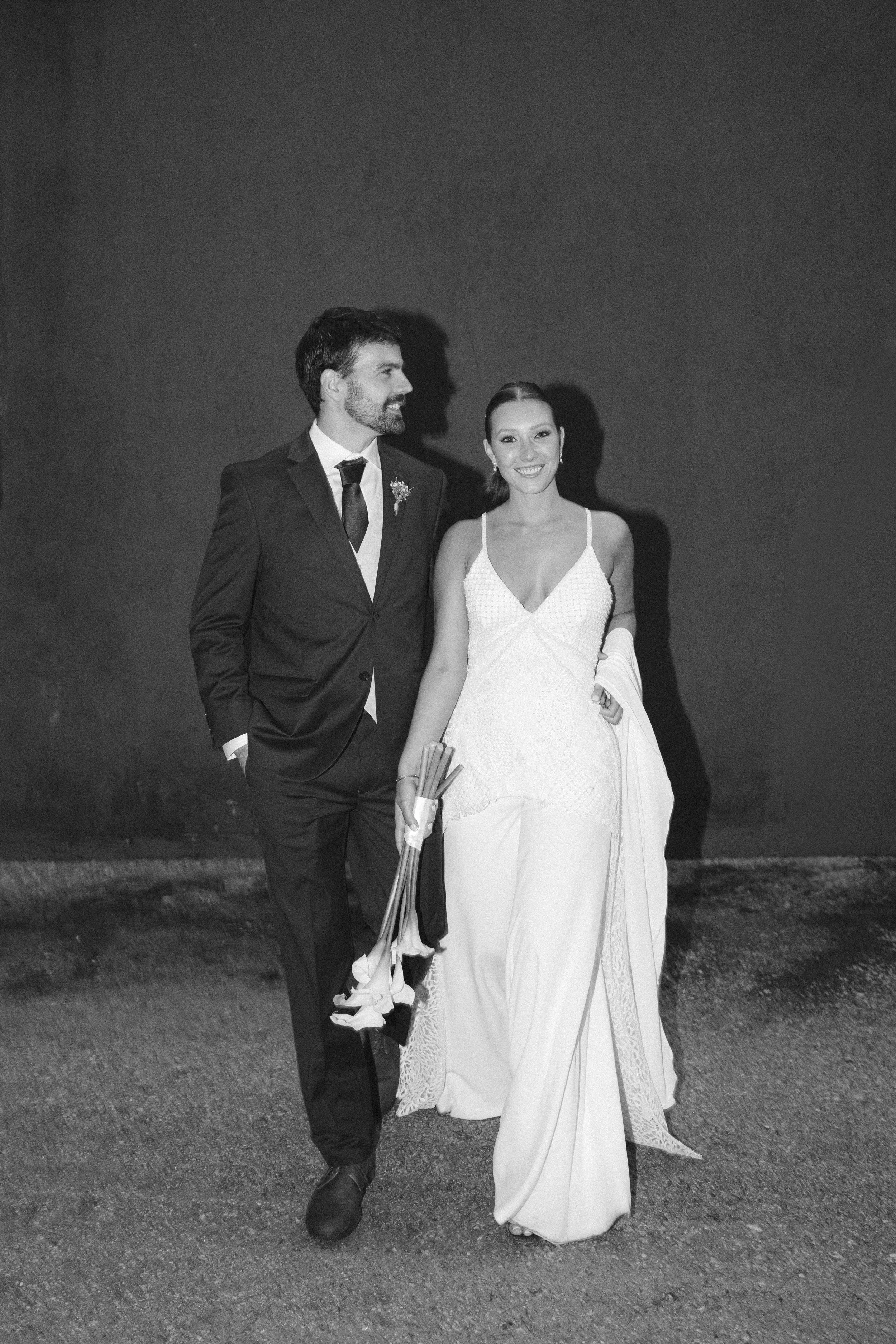Pareja en ropa formal, hombre con saco y mujer con vestido largo, caminando juntos y sonriendo. Ella lleva un ramo en la mano.