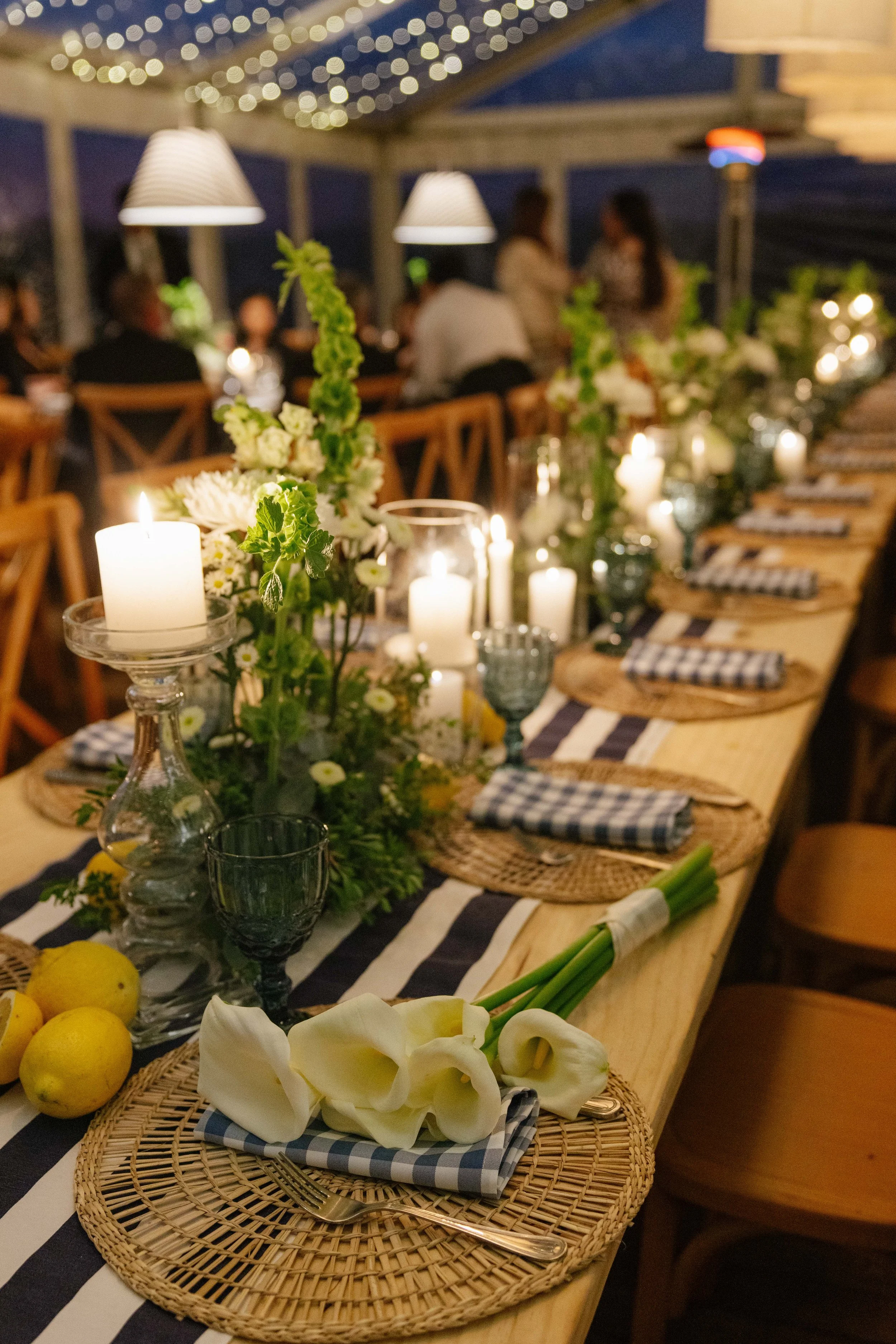 Mesa decorada con velas, flores blancas y verdes, limones, y platos con servilletas a cuadros. Ambiente cálido y elegante para una cena o evento.