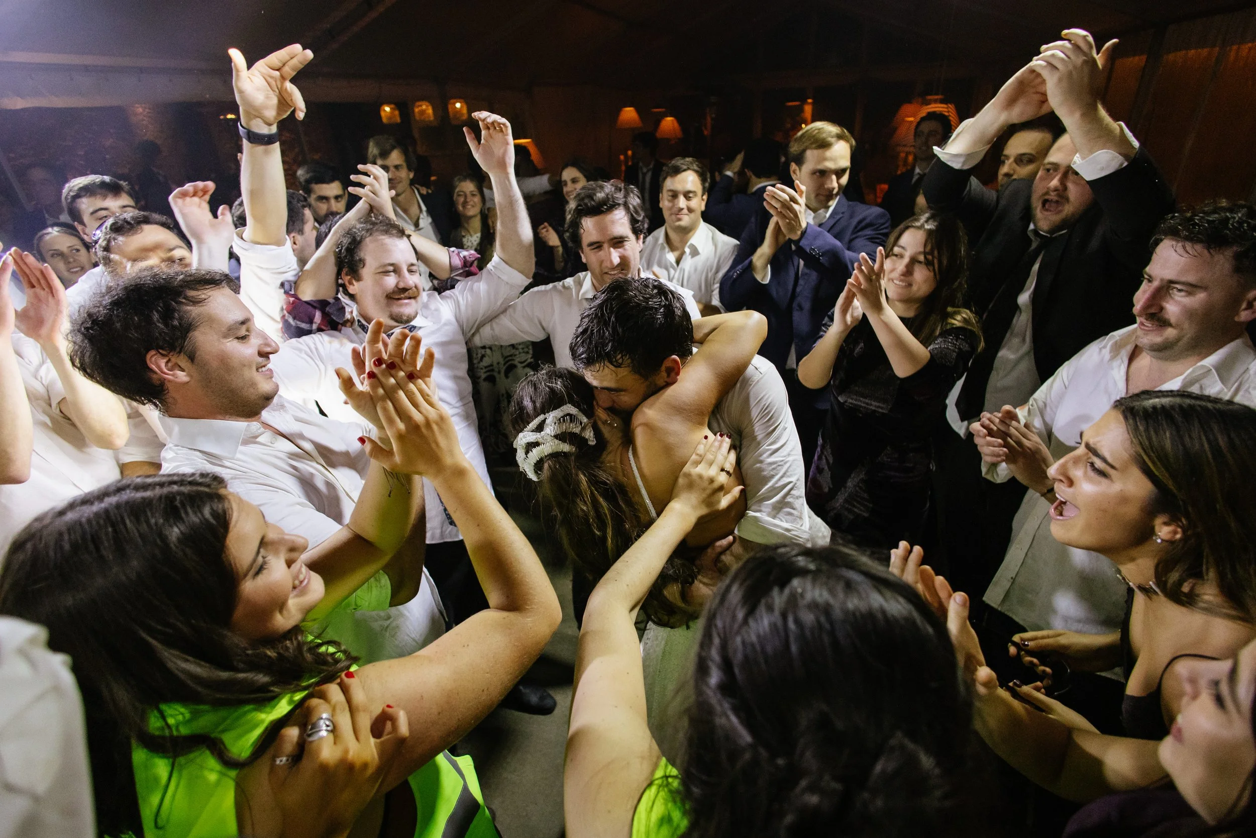 Grupo de personas bailando y disfrutando en una fiesta, algunos con sonrisas y otros abrazándose, en un ambiente animado con iluminación cálida.
