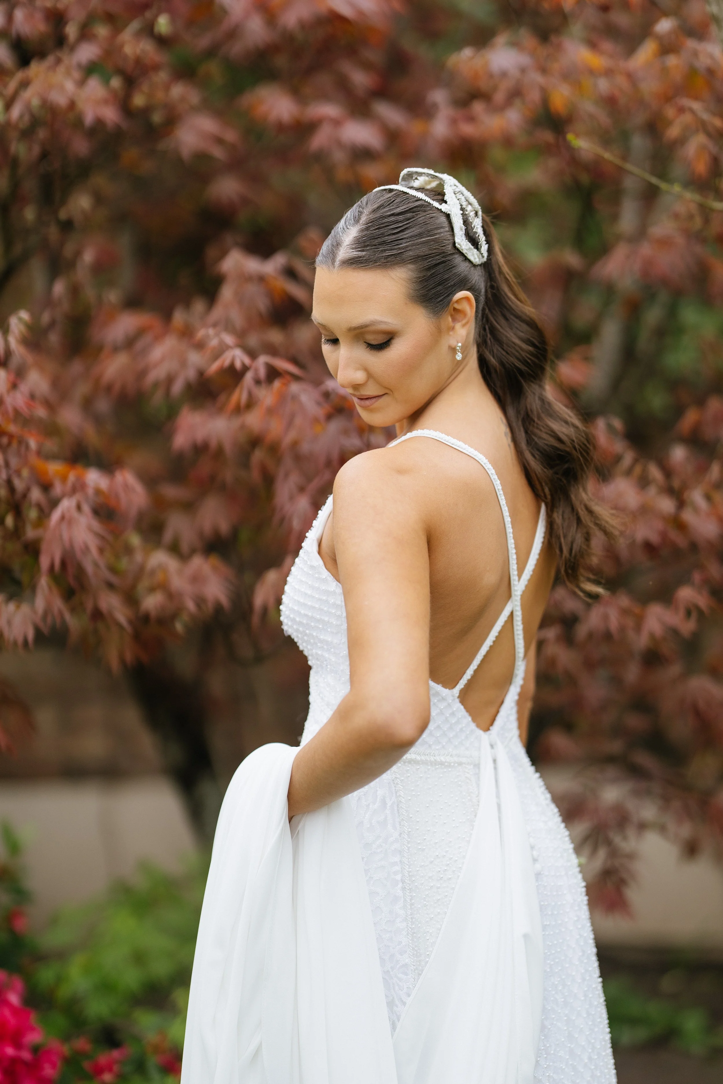 Mujer en vestido blanco con accesorios en el cabello, de cabello castaño oscuro y piel clara, en un entorno con hojas de color marrón y rojo.