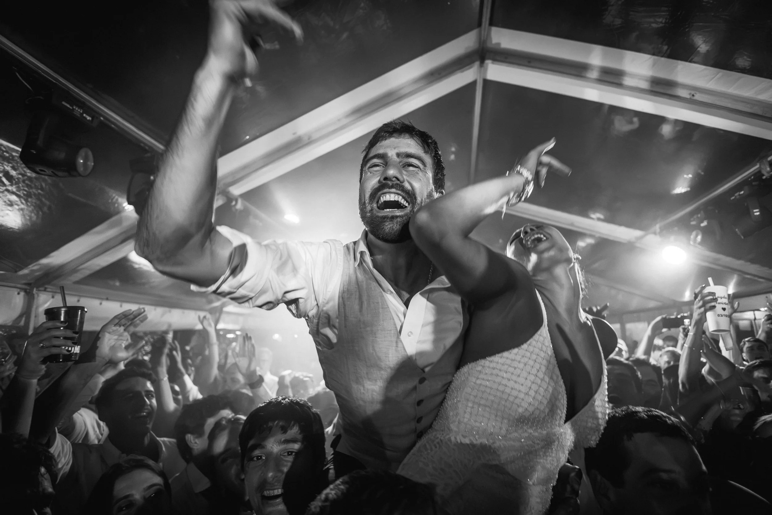Una pareja celebrando con muchas personas en un ambiente festivo, levantándose en un lugar con mucha luz y energía, en blanco y negro.
