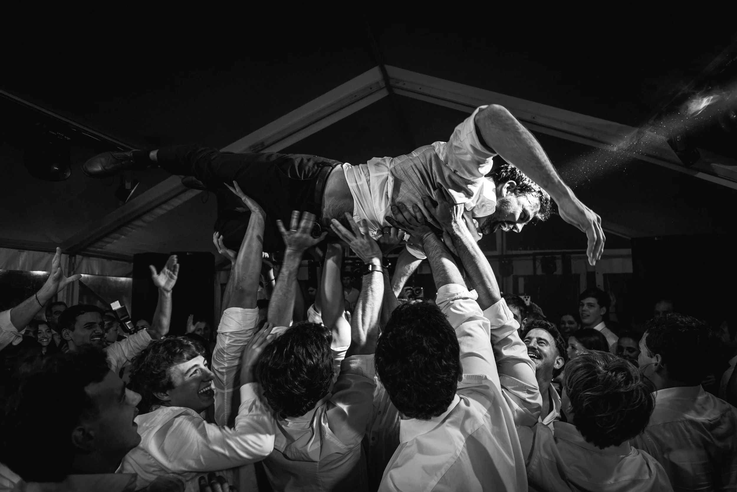 Hombre siendo levantado en una celebración por muchas manos en un lugar con techo de lona, en blanco y negro.