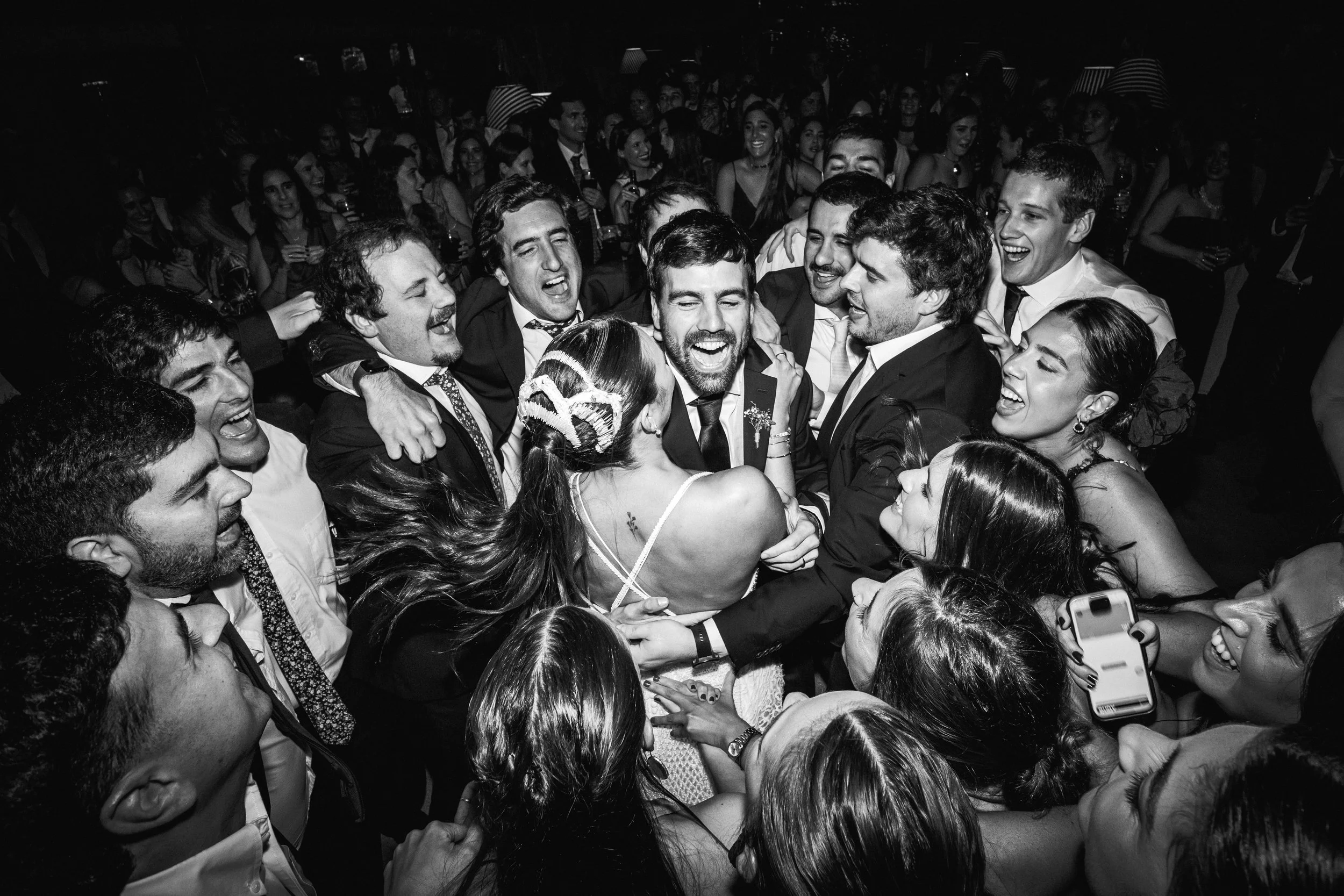 Grupo de personas en un evento social, riendo y abrazándose en un ambiente festivo, en blanco y negro.
