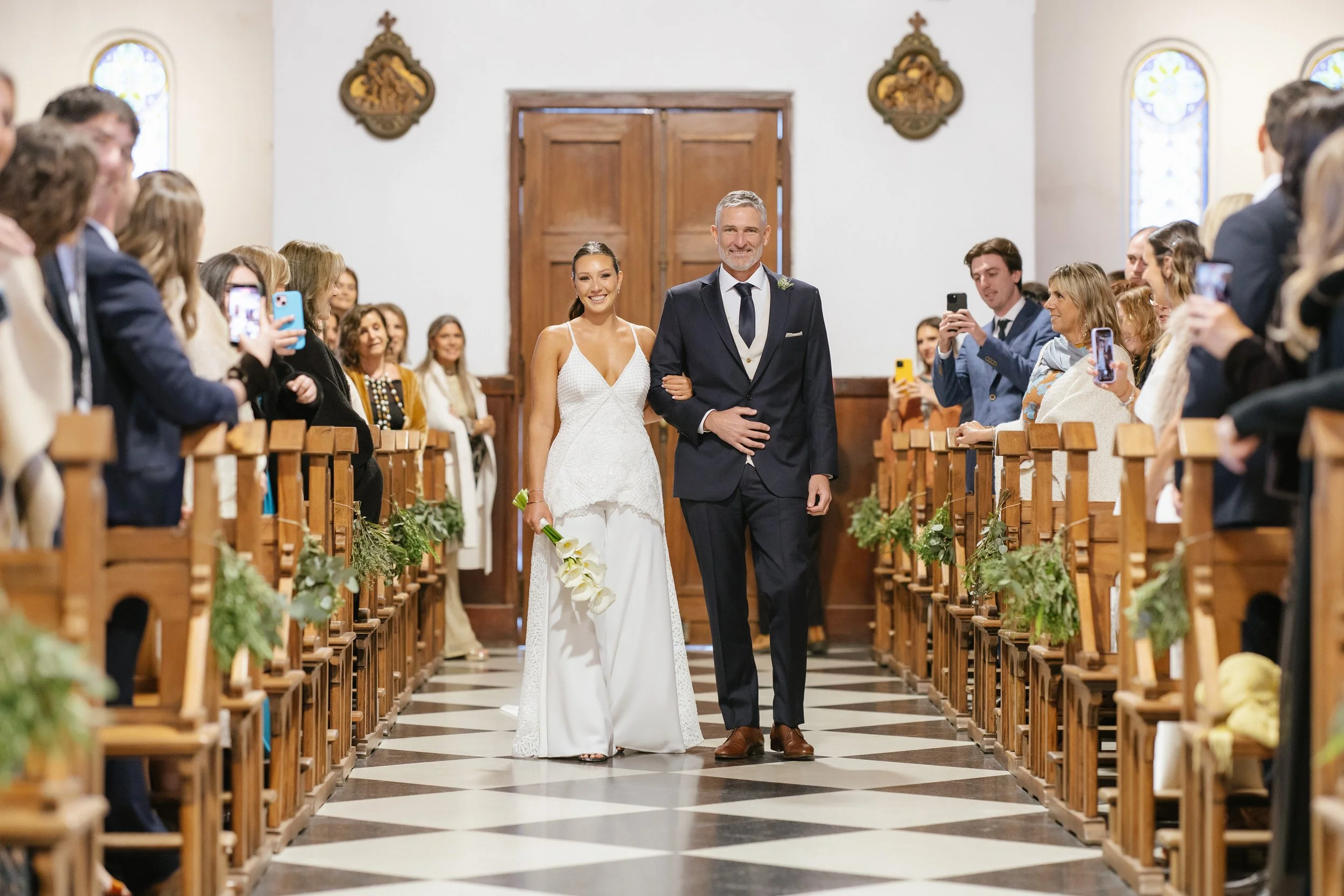 Pareja de novios caminando por la iglesia, rodeados de invitados que toman fotos, en una boda.