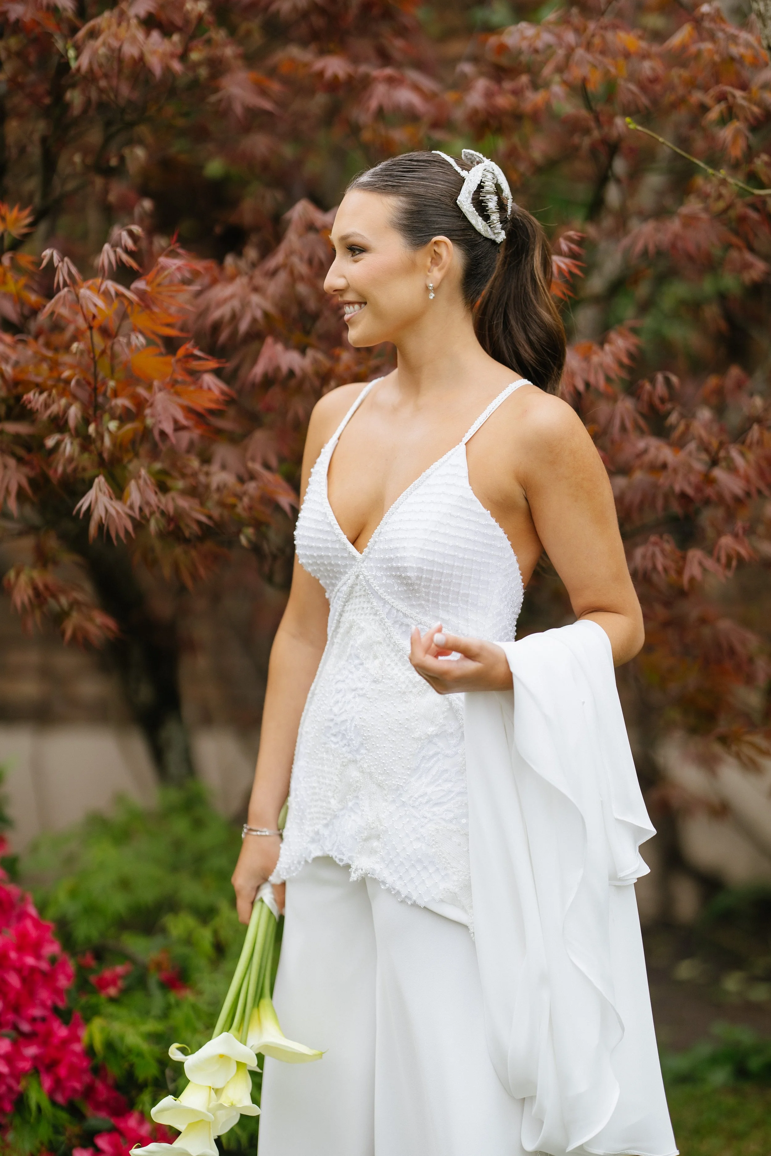 Una mujer sonriendo con vestido blanco de novia, sosteniendo un bouquet de flores blancas, en un fondo de hojas otoñales.