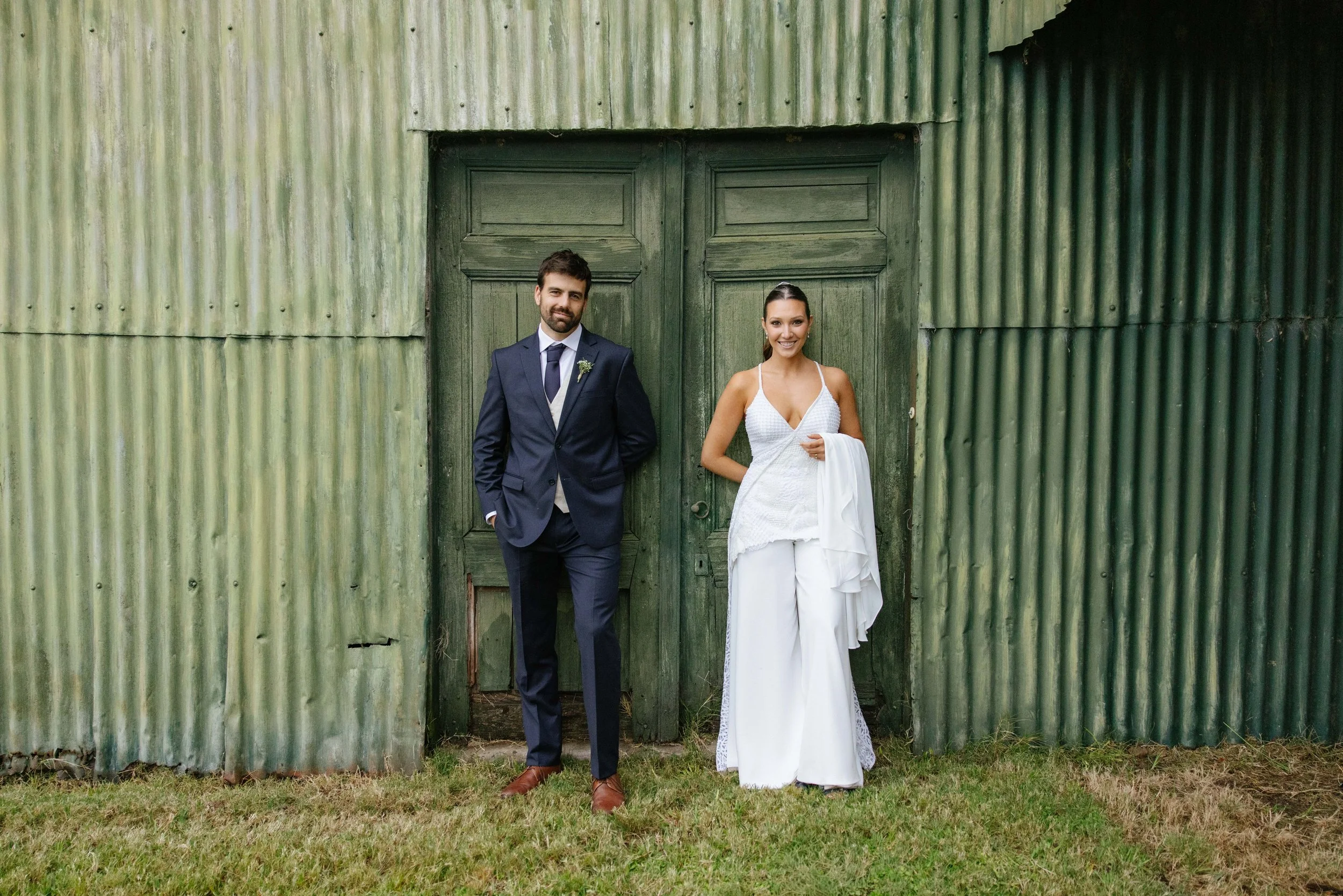 Pareja de boda posando frente a una puerta de madera en una estructura de láminas metálicas verdes, con el novio vestido con traje azul oscuro y la novia con vestido blanco, en un entorno rural.
