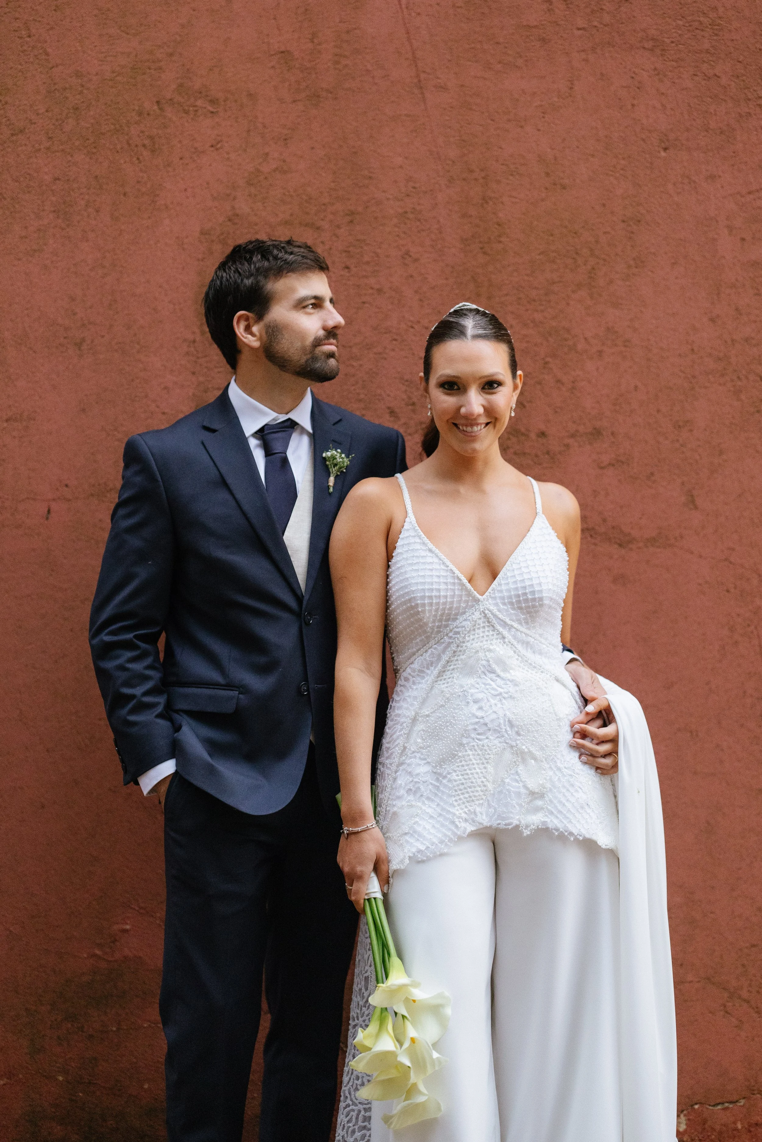 Pareja en vestido de boda, el hombre viste un traje azul oscuro con corbata y la mujer lleva un vestido blanco y sostiene un ramo de flores blancas, ambos posan frente a un muro rojo.