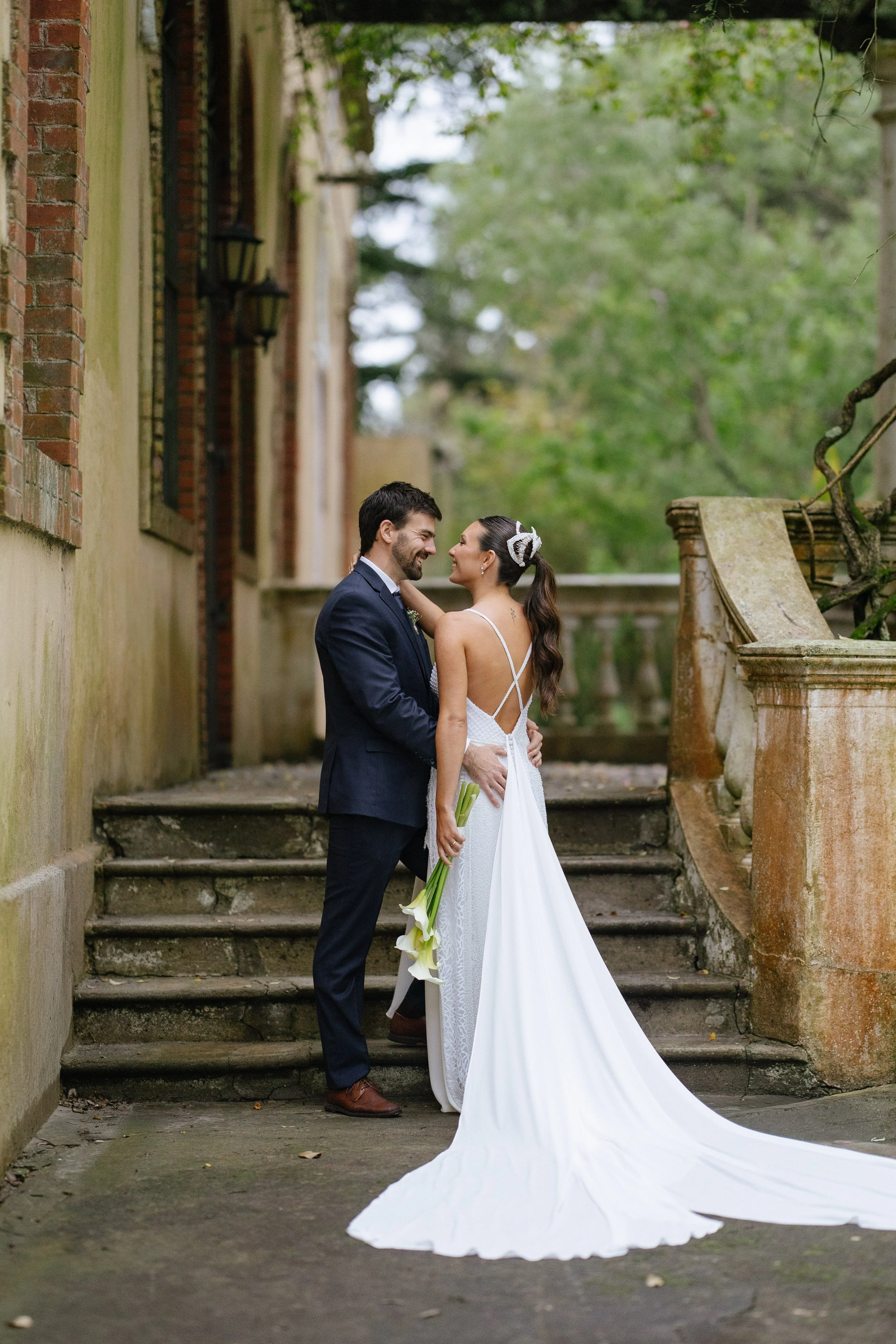 Una pareja de novios en una boda en un entorno al aire libre, con árboles y escaleras antiguas de piedra.