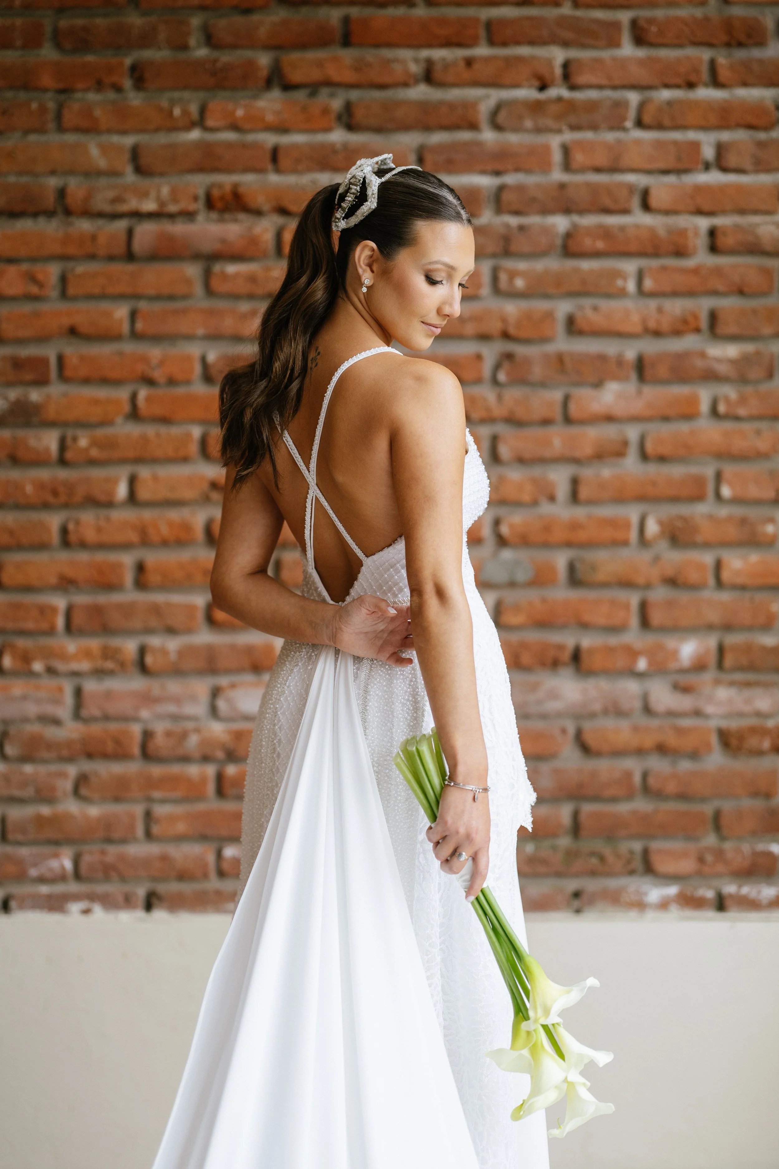 Mujer con vestido de novia en un interior con pared de ladrillos, sosteniendo un ramo de flores blancas.