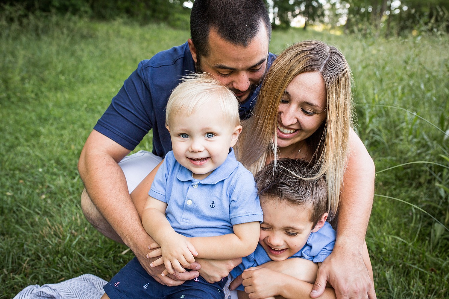  Photo of a mom, dad and their two boys hugging each other and giggling outside in the grass. 