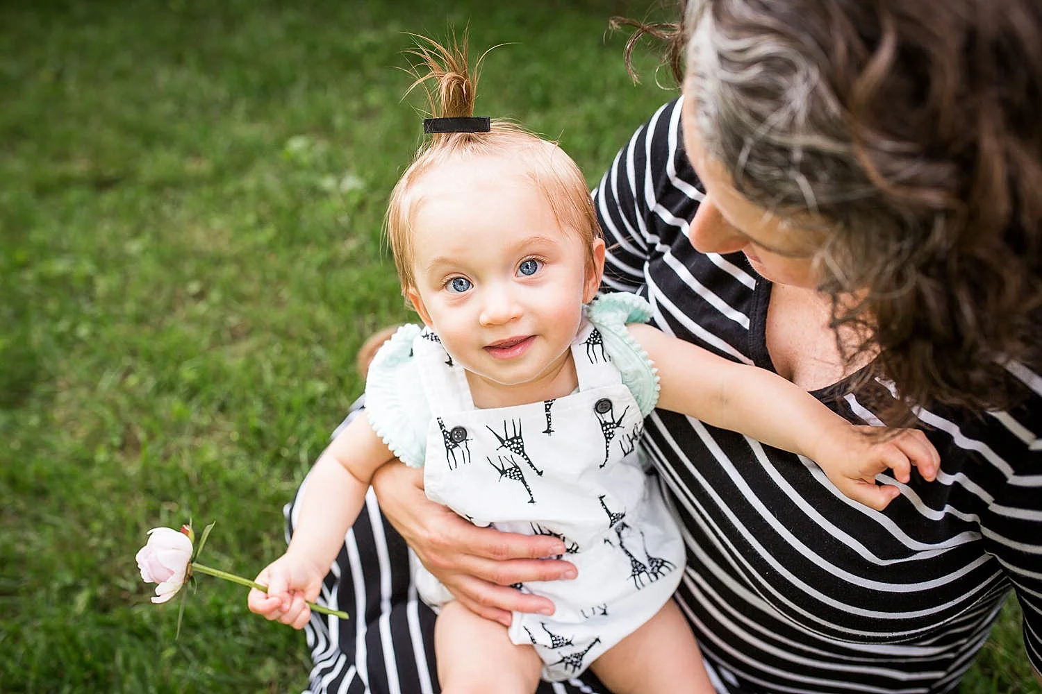 Photo of a little girl on her mother’s lap looking up and smiling with a flower in her hand.