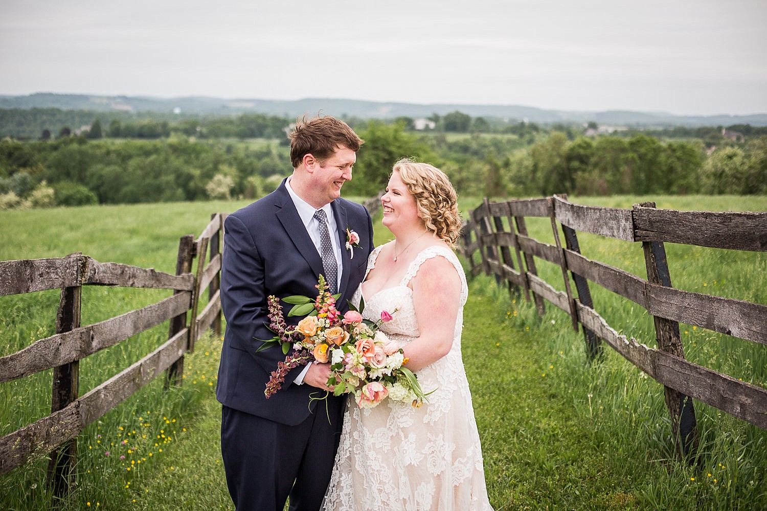  Photo of a bride and groom standing between a fence in a green field overlooking the Susquehanna River. 