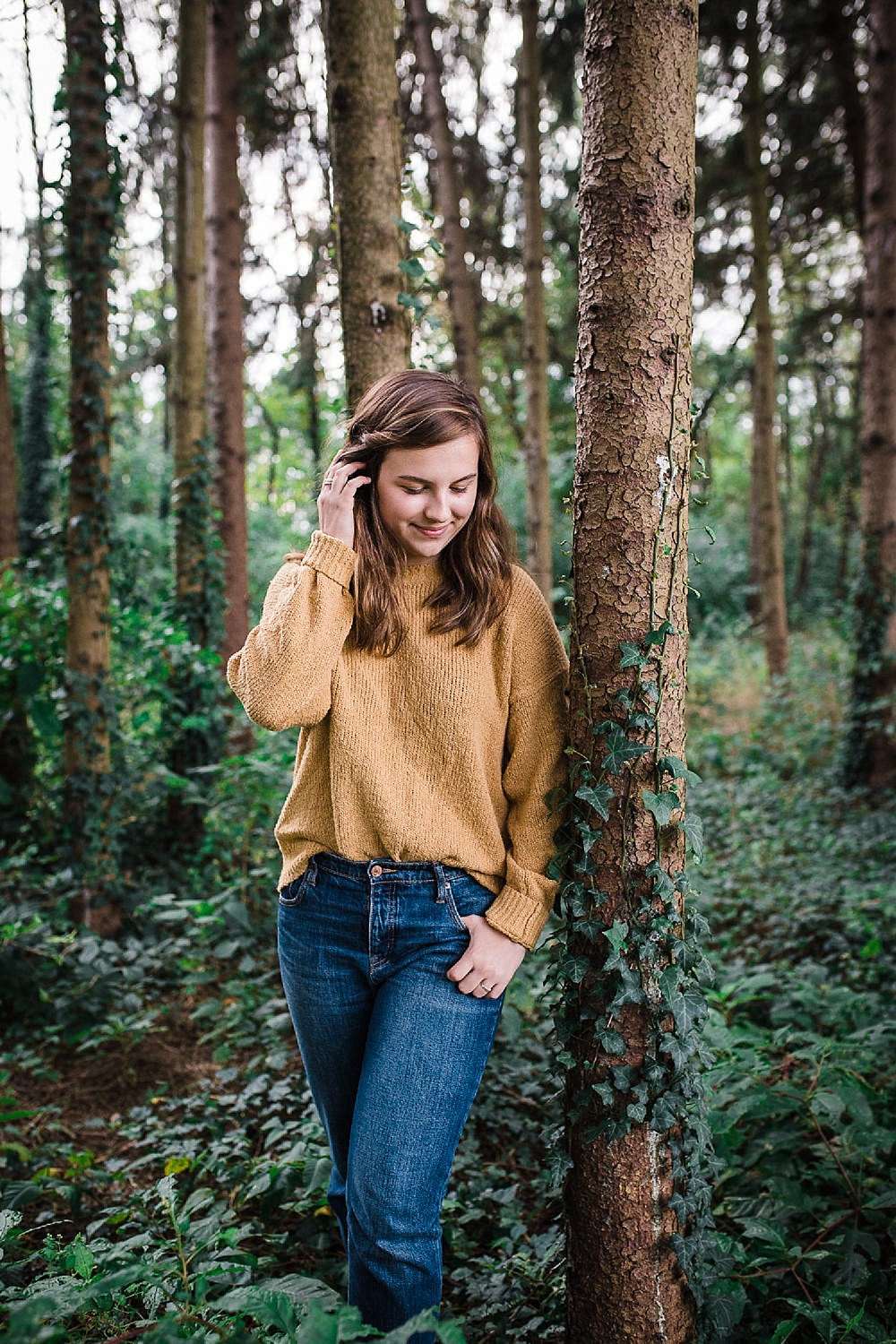  Lancaster senior session photo of a young girl in a yellow sweater and jeans standing in a pine forest. 