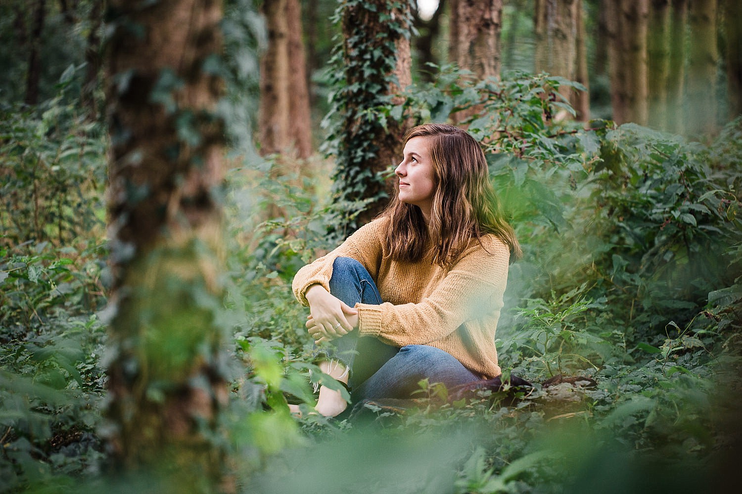  Lancaster senior session photo of a young girl in a yellow sweater and jeans sitting in a pine forest. 