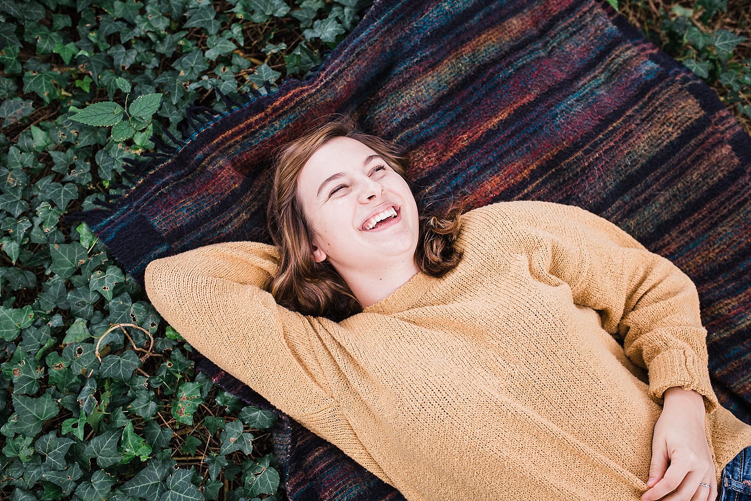  Lancaster senior session photo of a young girl in a yellow sweater and jeans lying on a striped blanket in a bed of ivy. 