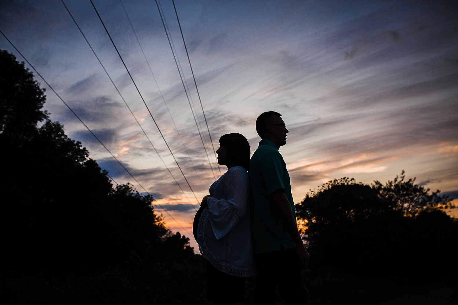  Sunset silhouette photo of a pregnant woman and a man standing back to back. 