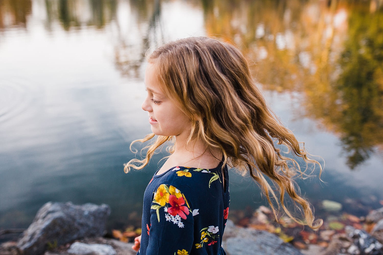Photo of a girl with long wavy hair standing in front of a pond with golden fall leaves in the reflection