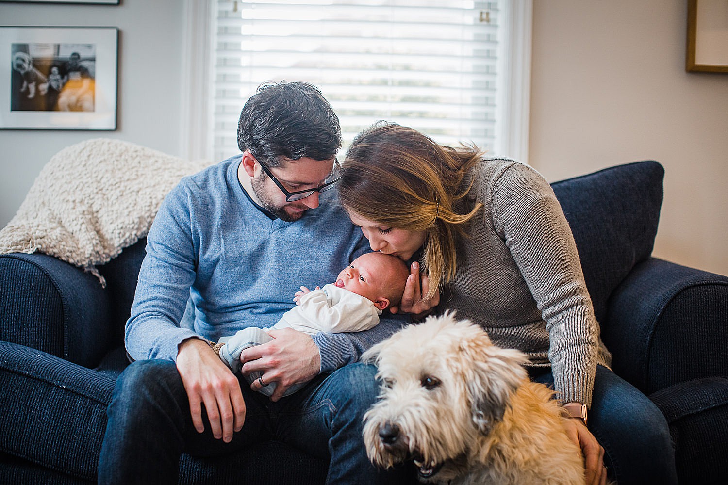  Photo of a mother and father holding their newborn son in their home with a shaggy dog in the foreground. 