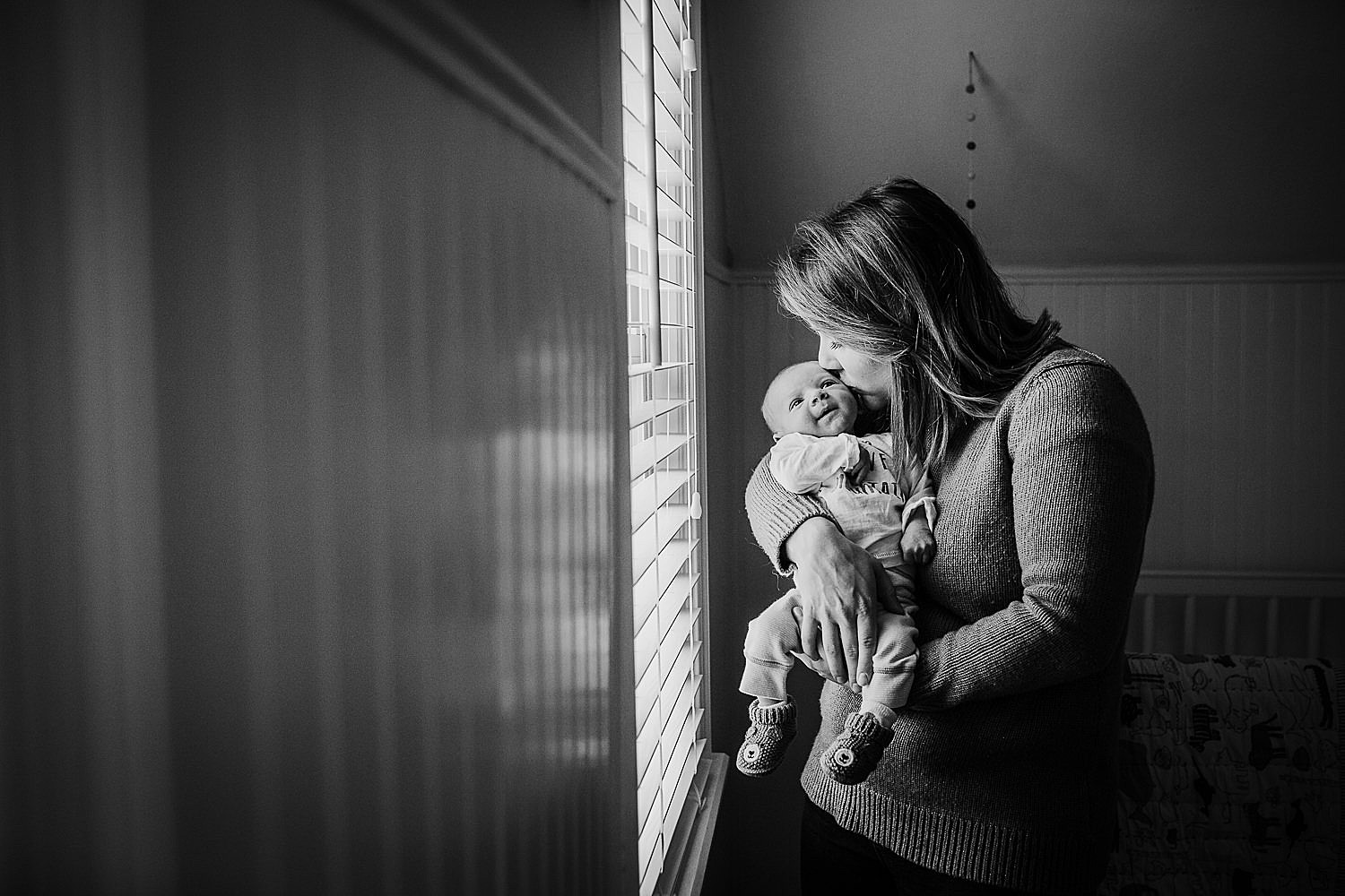  Photo of a mother kissing her newborn son in the nursery next to the window. 