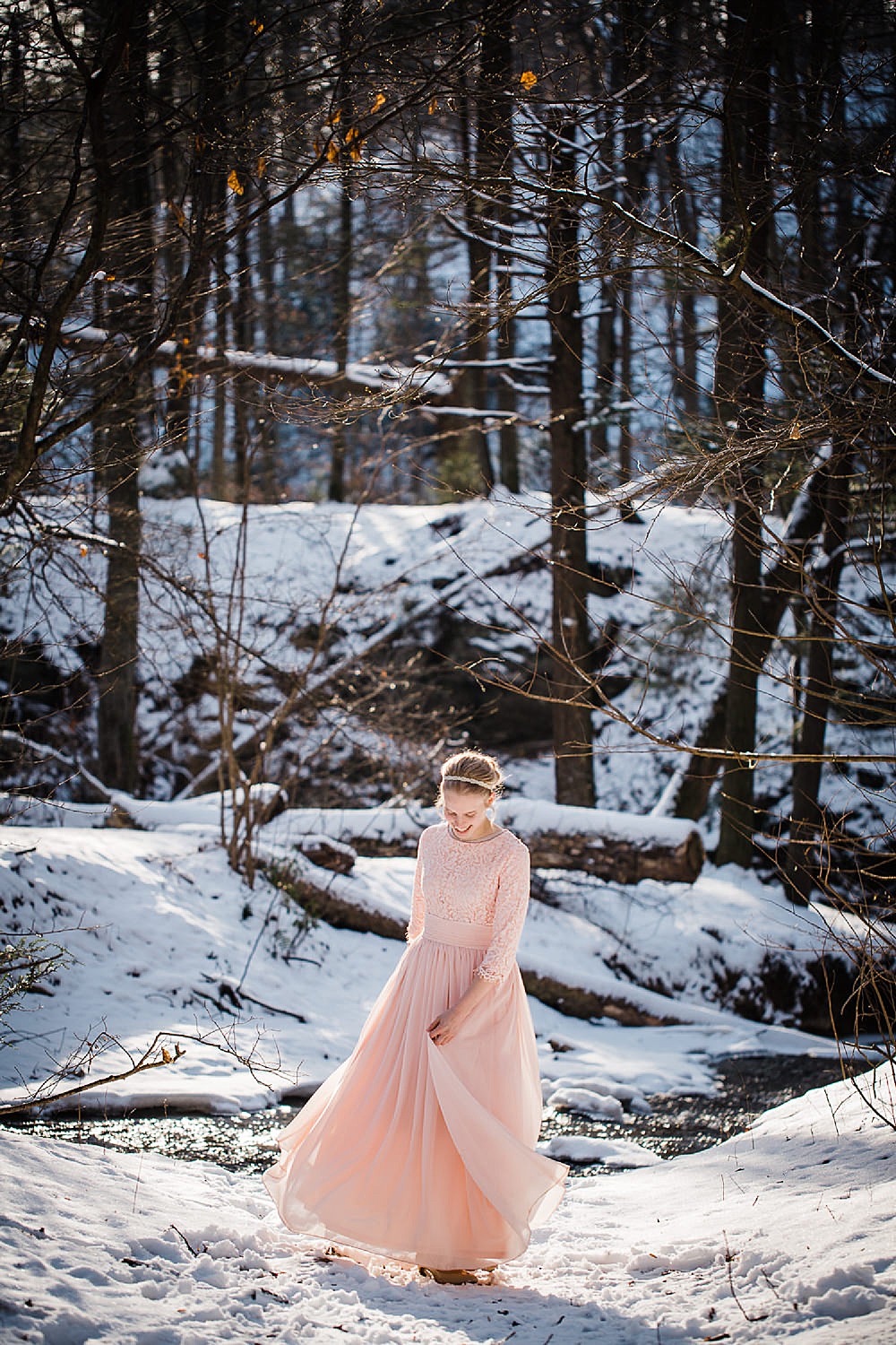  Photo of a young blonde woman in a long light pink gown twirling in a wintery forest. 
