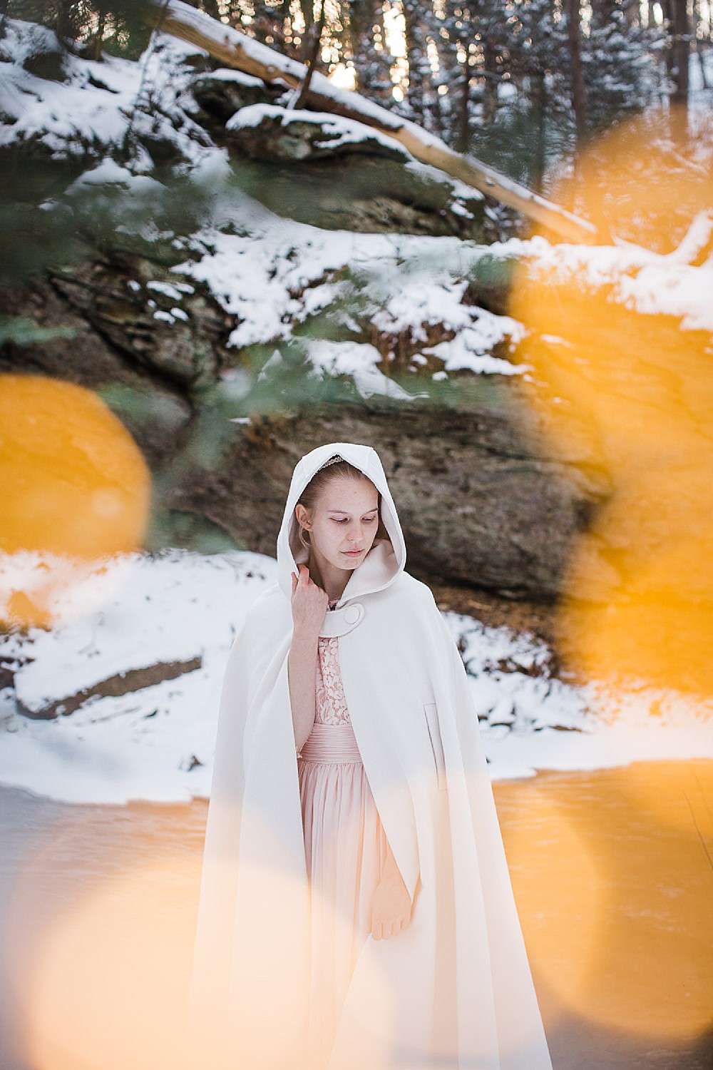  Photo of a young blonde woman in a long light pink gown and white hooded cape standing in a wintery forest. 