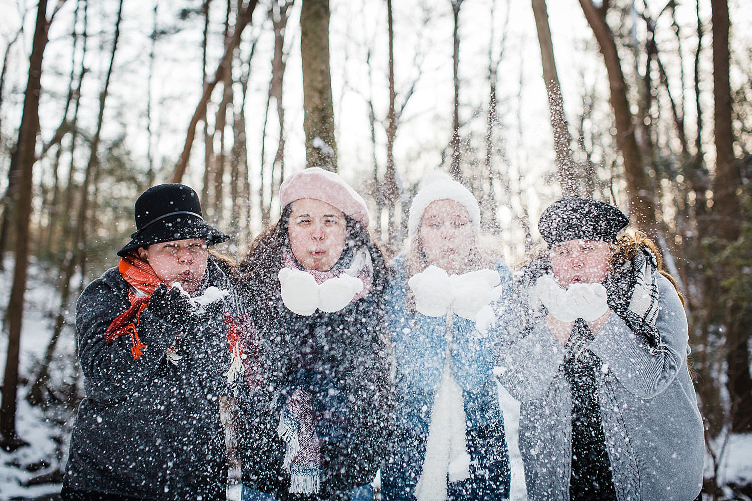  Photo of four young women blowing snow in the air in a wintery forest. 