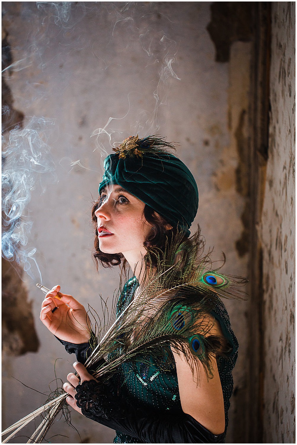  Photo of a glamorous young brunette woman wearing a green vintage flapper dress and a green velvet turban and peacock feathers holding a smoking cigarette in her hand. 