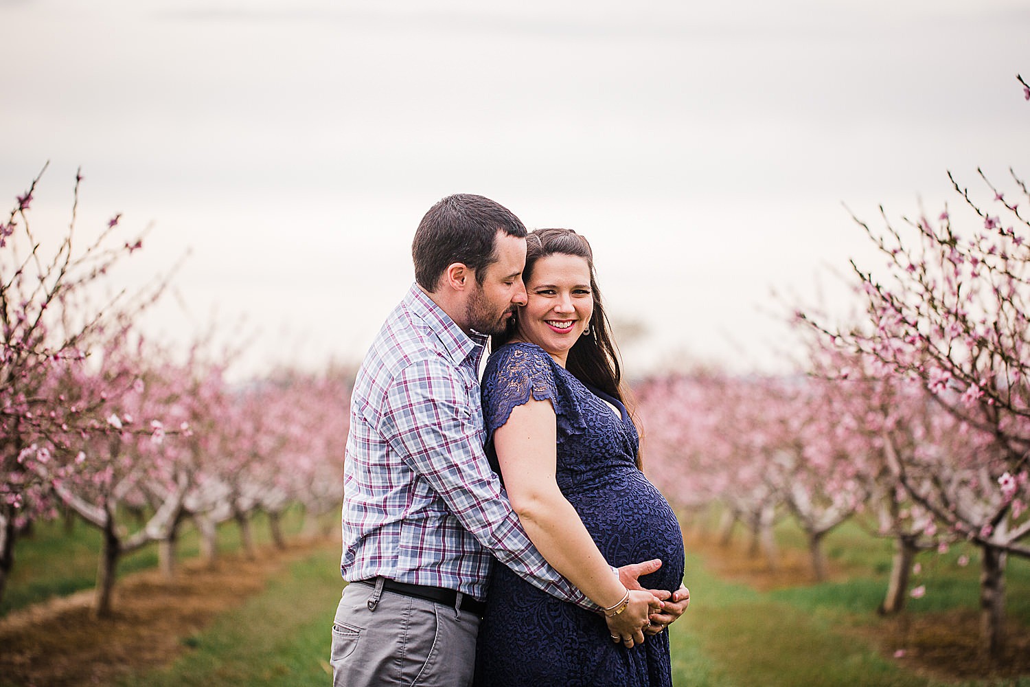 Photo of a pregnant woman in a blue lace dress standing with her husband in an orchard of flowers.