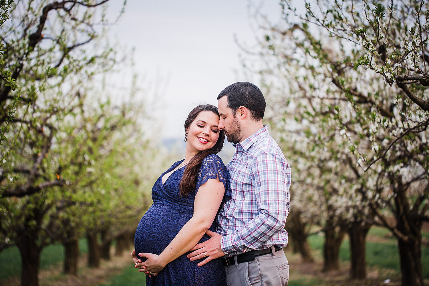 Photo of a pregnant woman in a blue lace dress standing with her husband in an orchard of flowers.