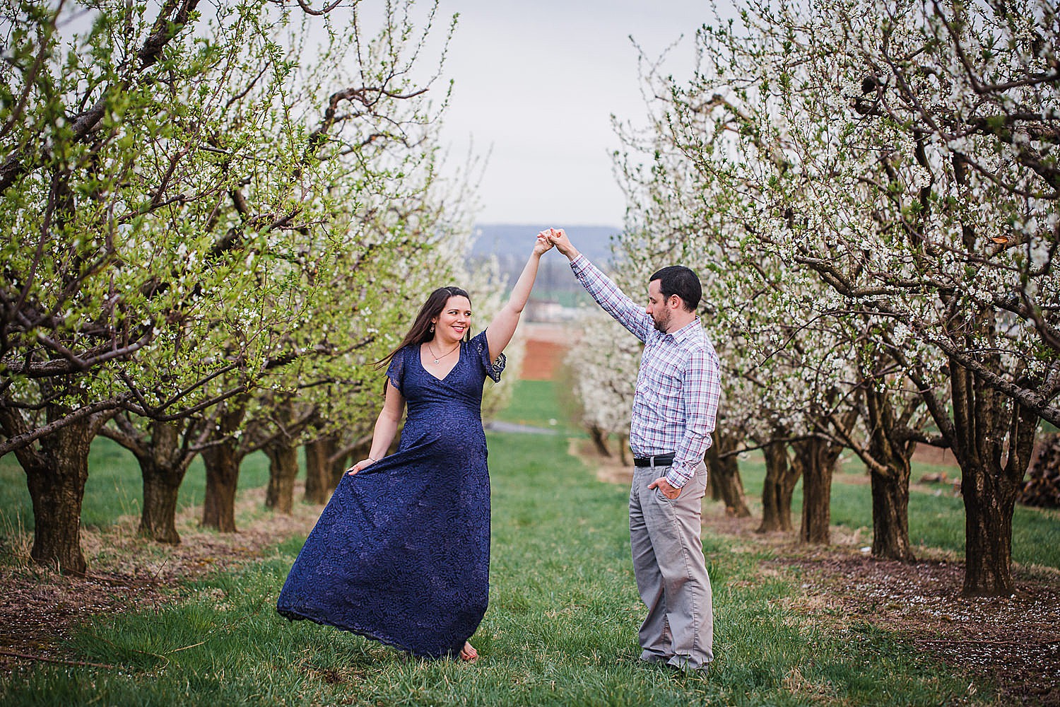  Photo of a pregnant woman in a blue lace dress dancing with her husband in an orchard of flowers. 