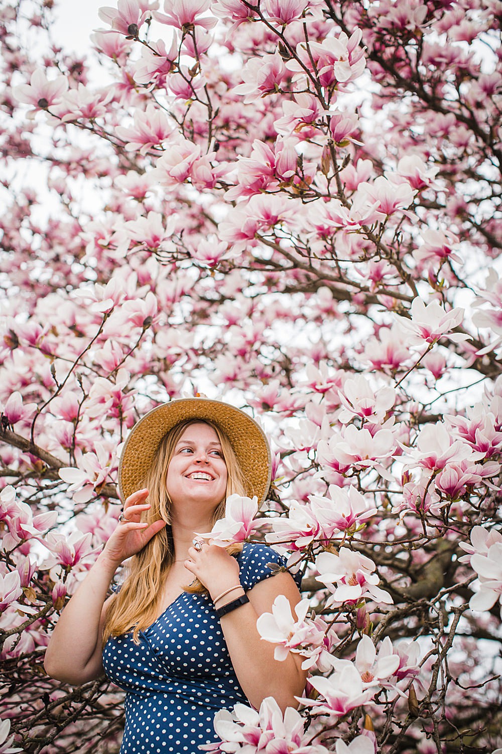  Photo of a young woman in a straw hat and polka dot dress standing in a tree of magnolia blossoms. 